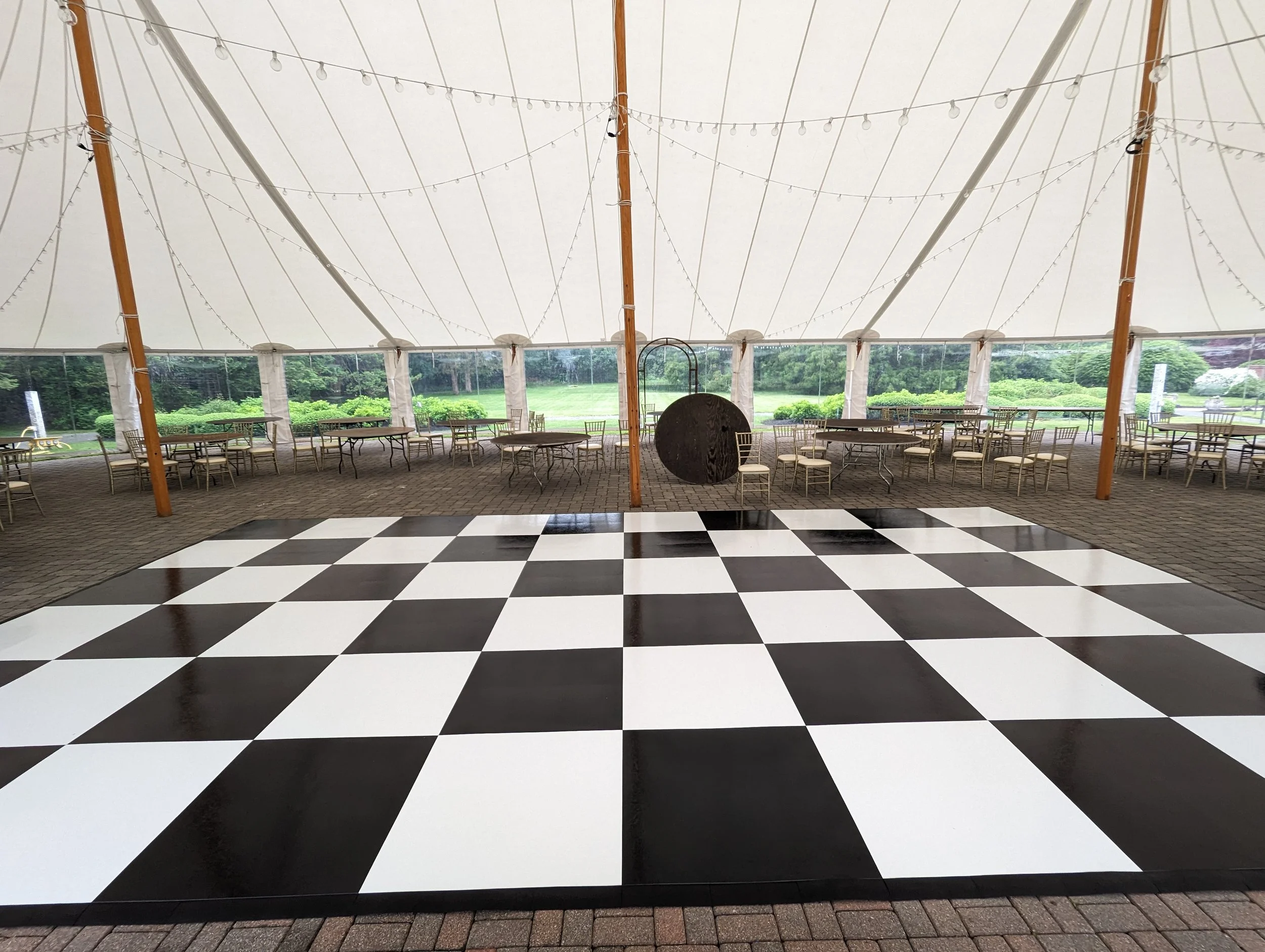 Interior of a large event tent with a black and white checkered dance floor and a seating area with tables and chairs along the sides, decorated with string lights.