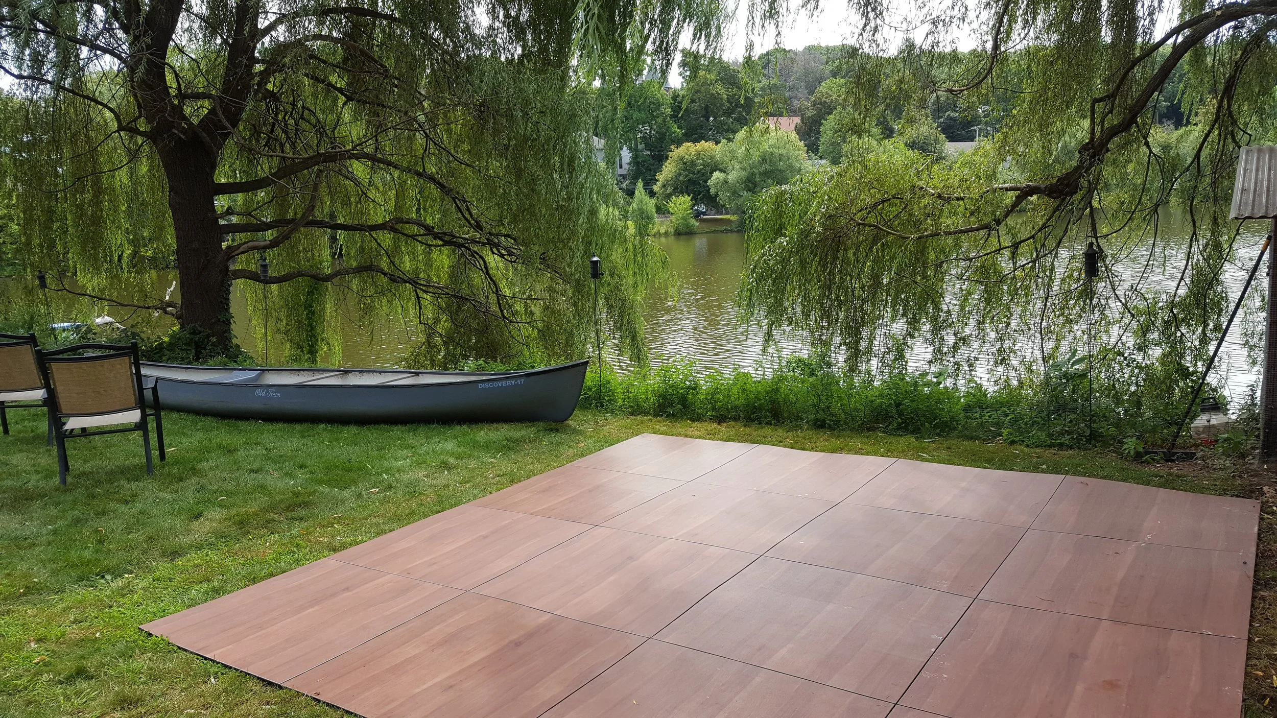 A lakeside scene with a weeping willow tree, a boat resting on the grass, and a small tiled patio area.