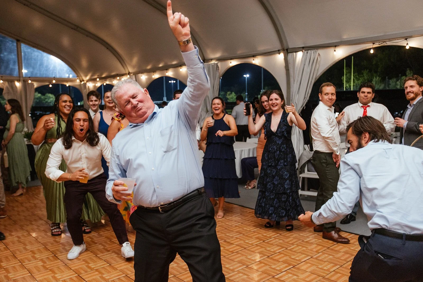 People dancing and enjoying themselves at a party under a large tent with string lights.