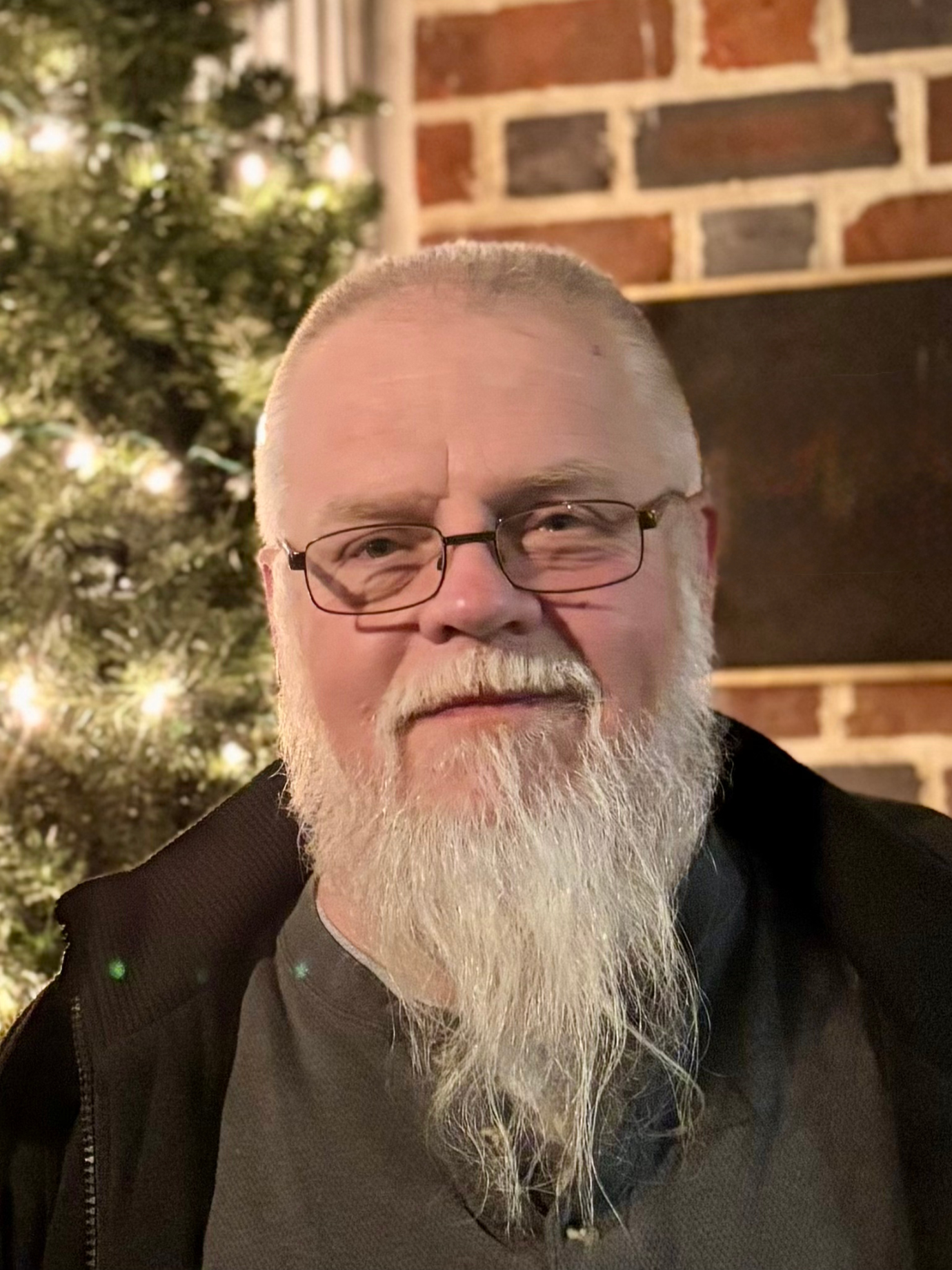 A man with glasses and a long white beard, smiling, in front of a decorated Christmas tree and a brick fireplace.