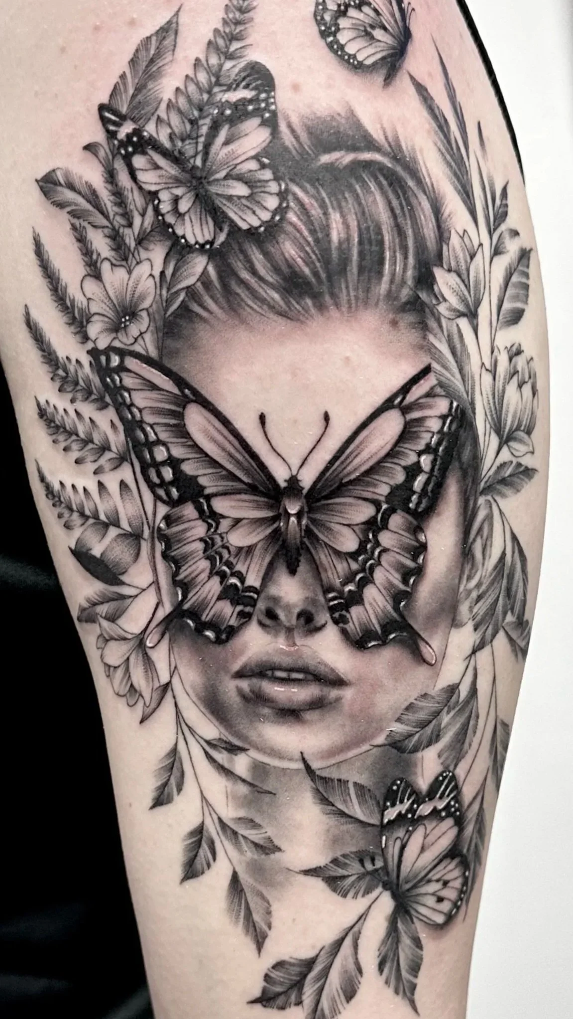 Black and grey tattoo of a woman's face with butterflies and leaves surrounding it.