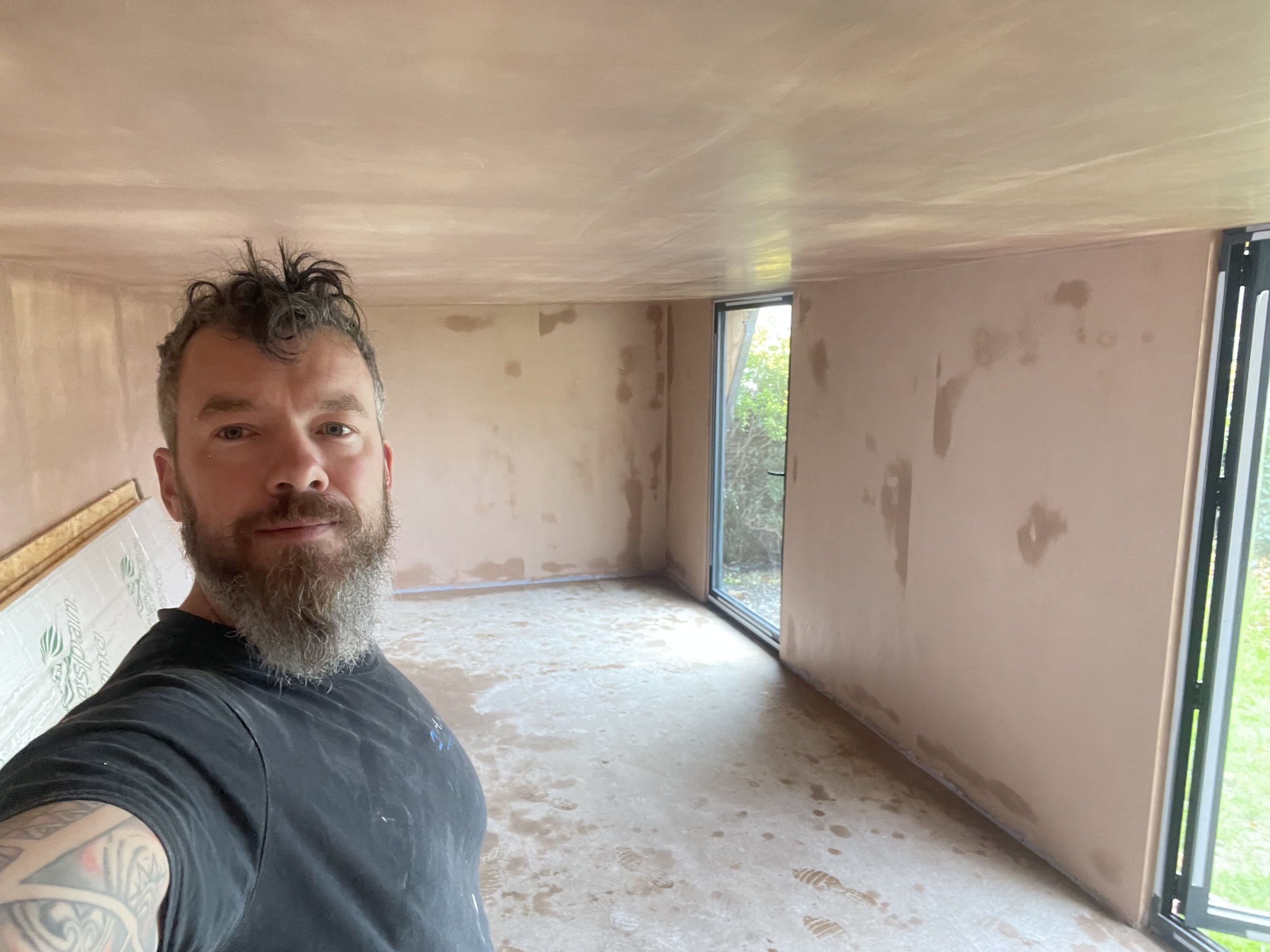 Man with a beard taking a selfie inside a room under construction with unfinished walls and large windows.