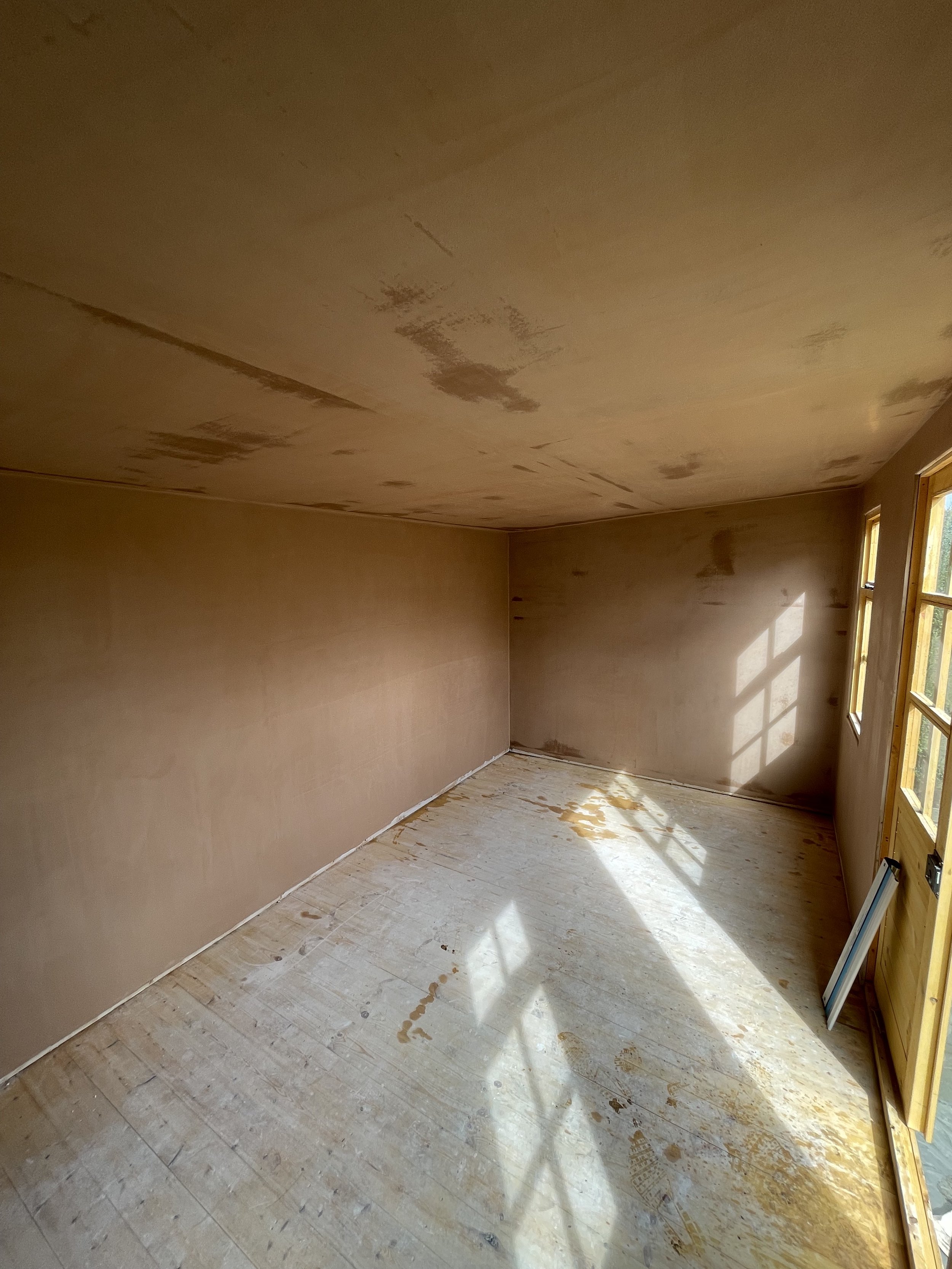 Empty room under renovation with unfinished walls and floor, sunlight coming through a window, and a door on the right side.