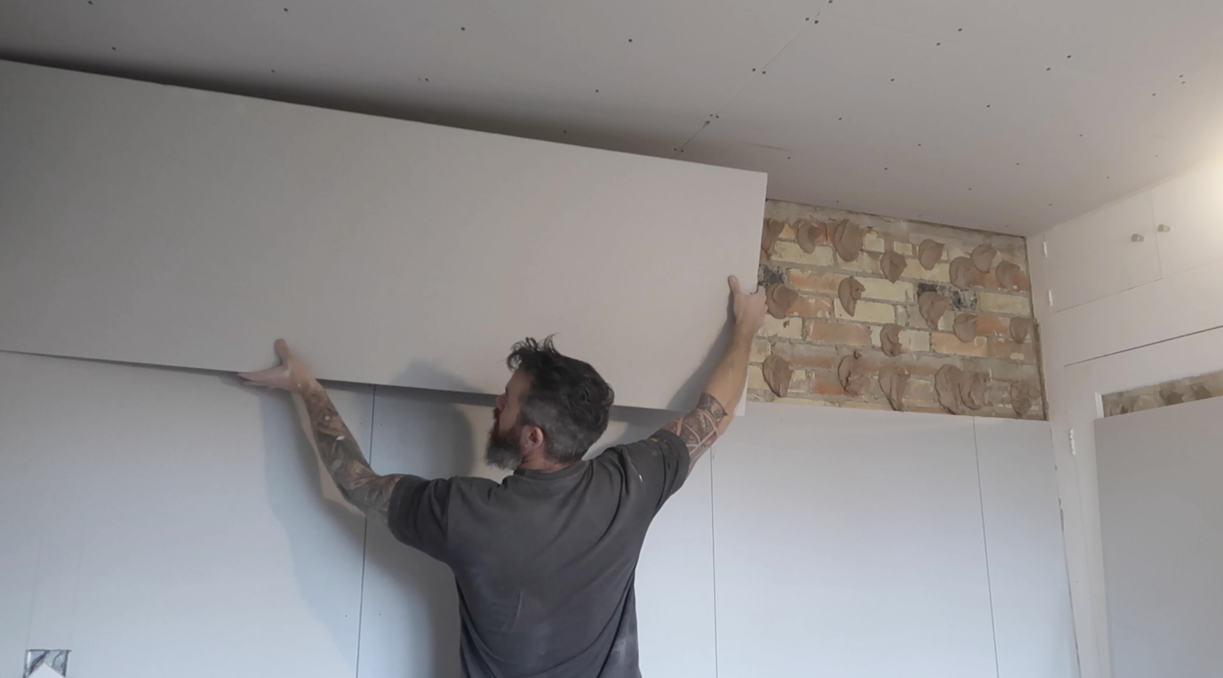A man with tattoos on his arms installing a large white drywall panel on a wall in a room under construction, with exposed bricks and partially finished ceiling.
