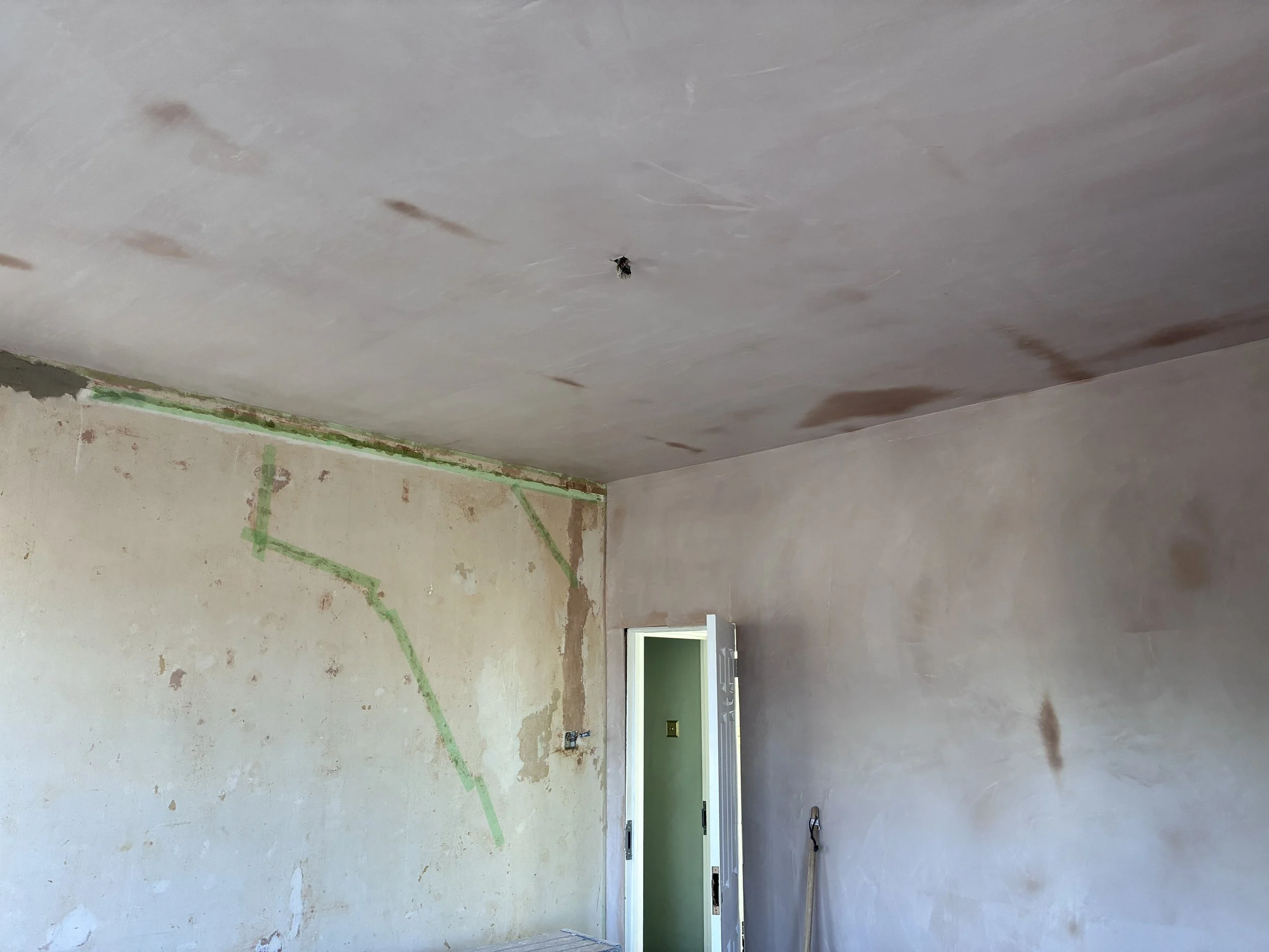 Interior of a room under renovation with unfinished walls and ceiling, with green painter's tape marking areas on the wall, and an open door leading to another room.
