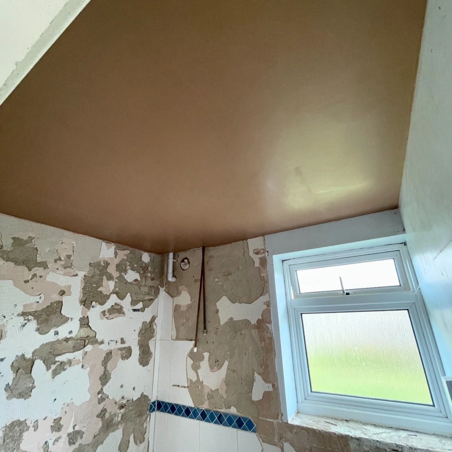 A room under construction with peeling wall paint, a partially finished ceiling, exposed pipes and wires, a window with bamboo shade, and interior wall in progress.