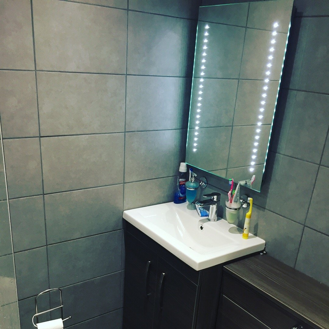 Bathroom with a rectangular mirror with LED lights, a white sink, and a dark vanity cabinet. Items on the sink including toothbrushes, toothpaste, soap dispenser, and cleaning products. Tiled wall and a toilet paper holder on the left wall.