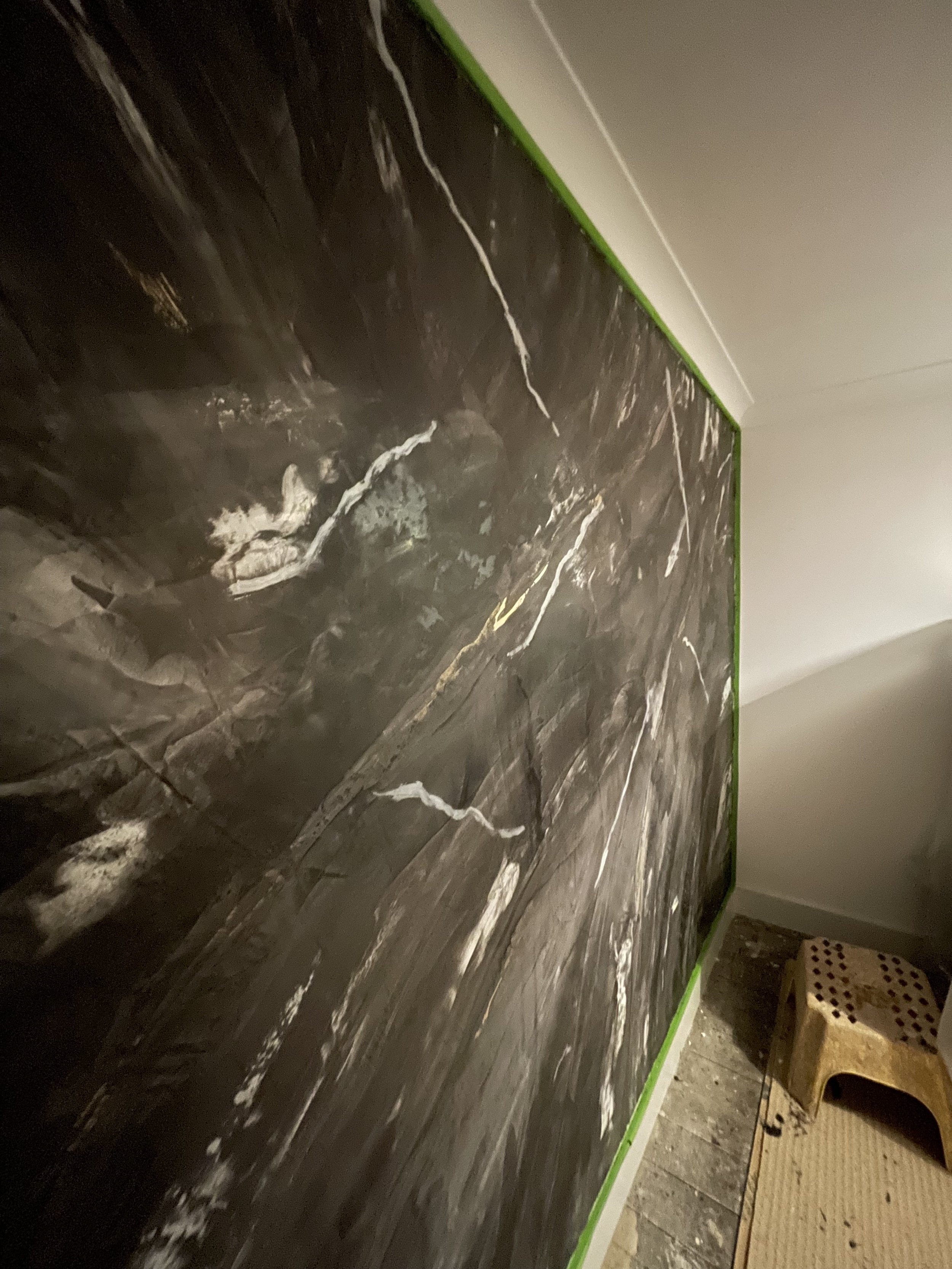 A close-up of a dark marble wall with white veining, partially bordered with green painter's tape, next to a small wooden stool on a wooden floor.
