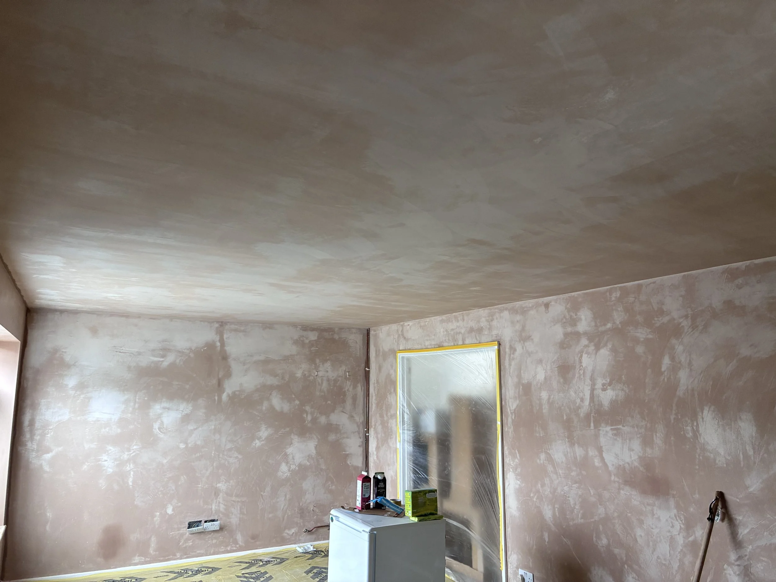 Room under renovation with unpainted beige walls, plastic covering on the floor, partially covered window, and various items such as bottles, cans, and a white cabinet on the floor.