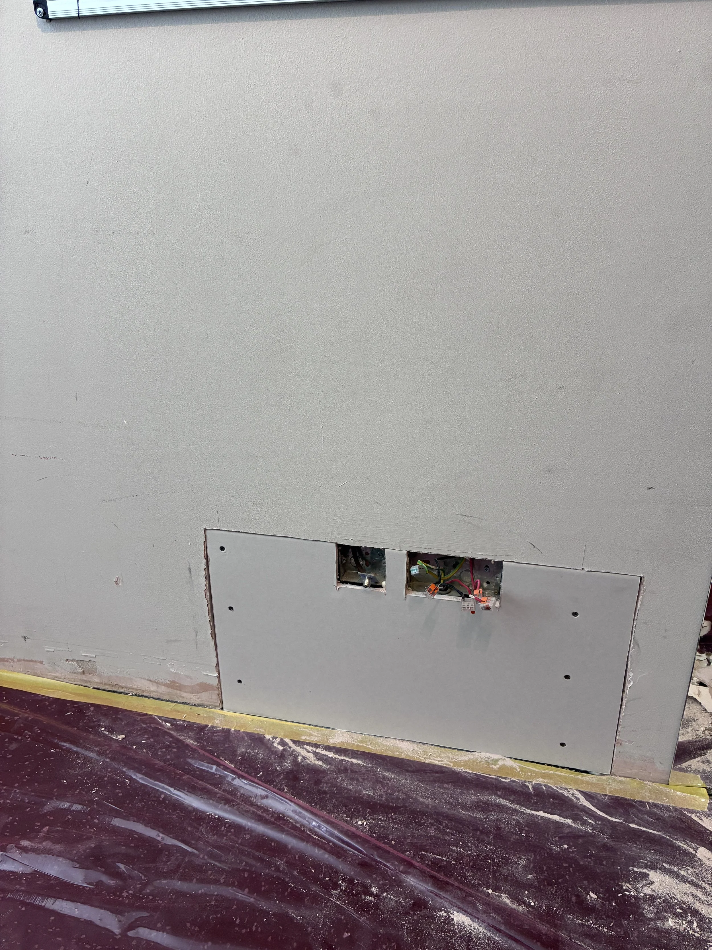 Unfinished drywall with an electrical box and wires, with a plastic sheet on the floor to collect debris.