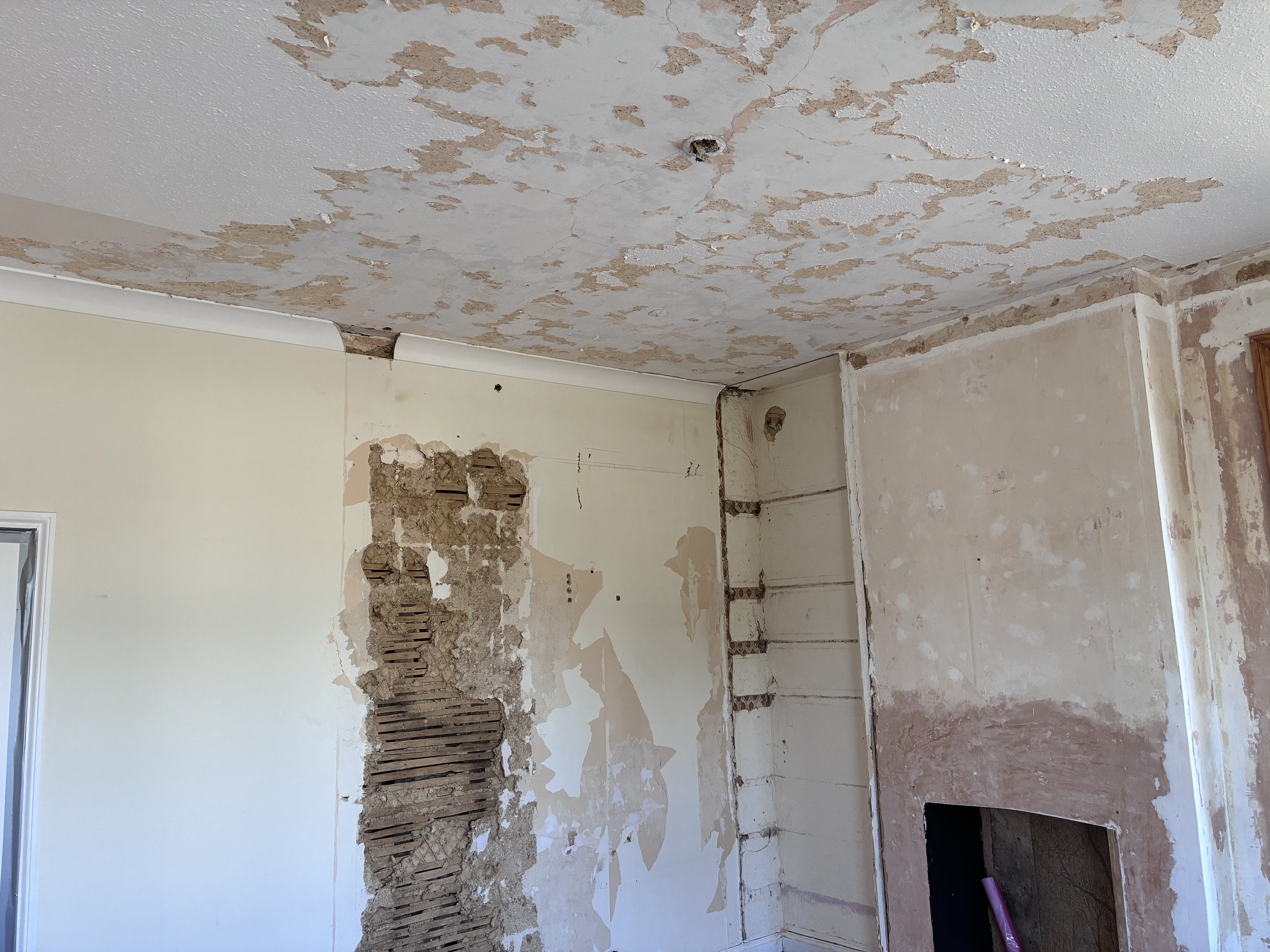 A room undergoing renovation with peeling paint on the ceiling and wall. The ceiling has many patches where paint is chipped off, exposing the drywall. The wall has similar damage with holes and missing plaster, revealing underlying materials. There is a small window on the left side and a pile of construction tools on the floor.