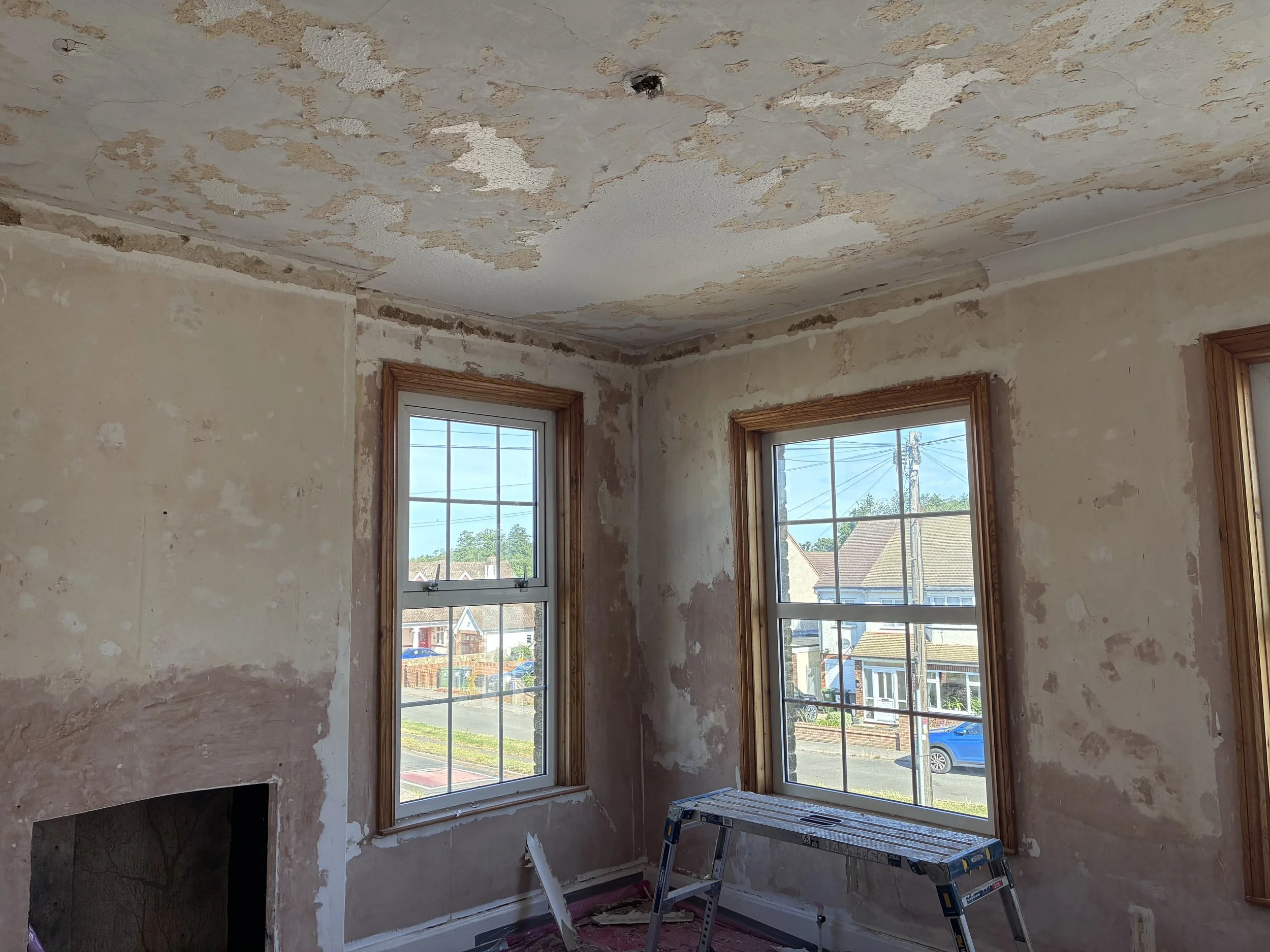 A room under renovation with peeling paint or wallpaper on the ceiling and walls. Two windows with wooden trim allow natural light inside, revealing a view of neighboring houses and a street outside. A step ladder is in the foreground, indicating ongoing work.