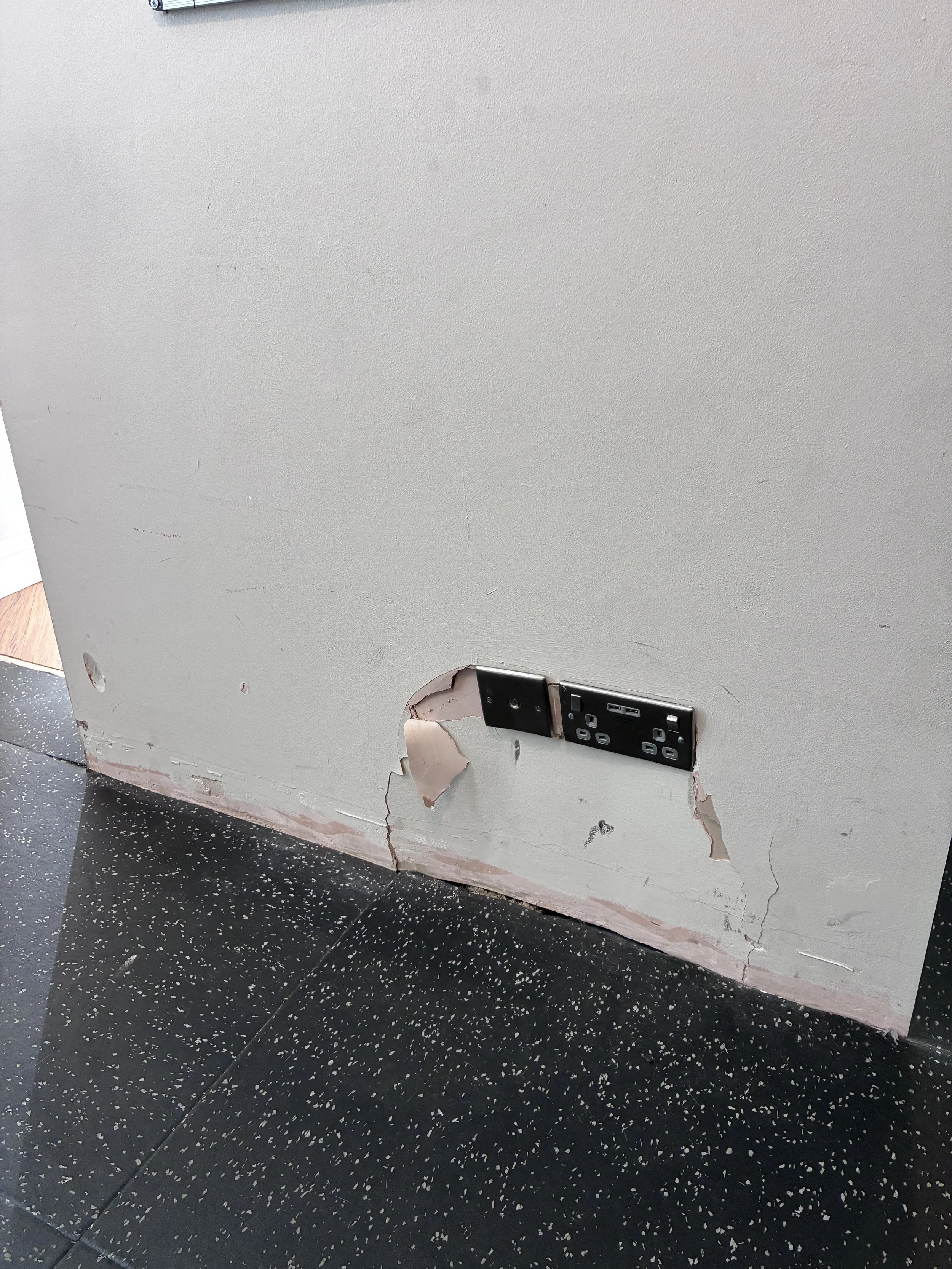 The wall with an electrical outlet, visible damages, and peeling paint near the bottom. The flooring is black with white specks.