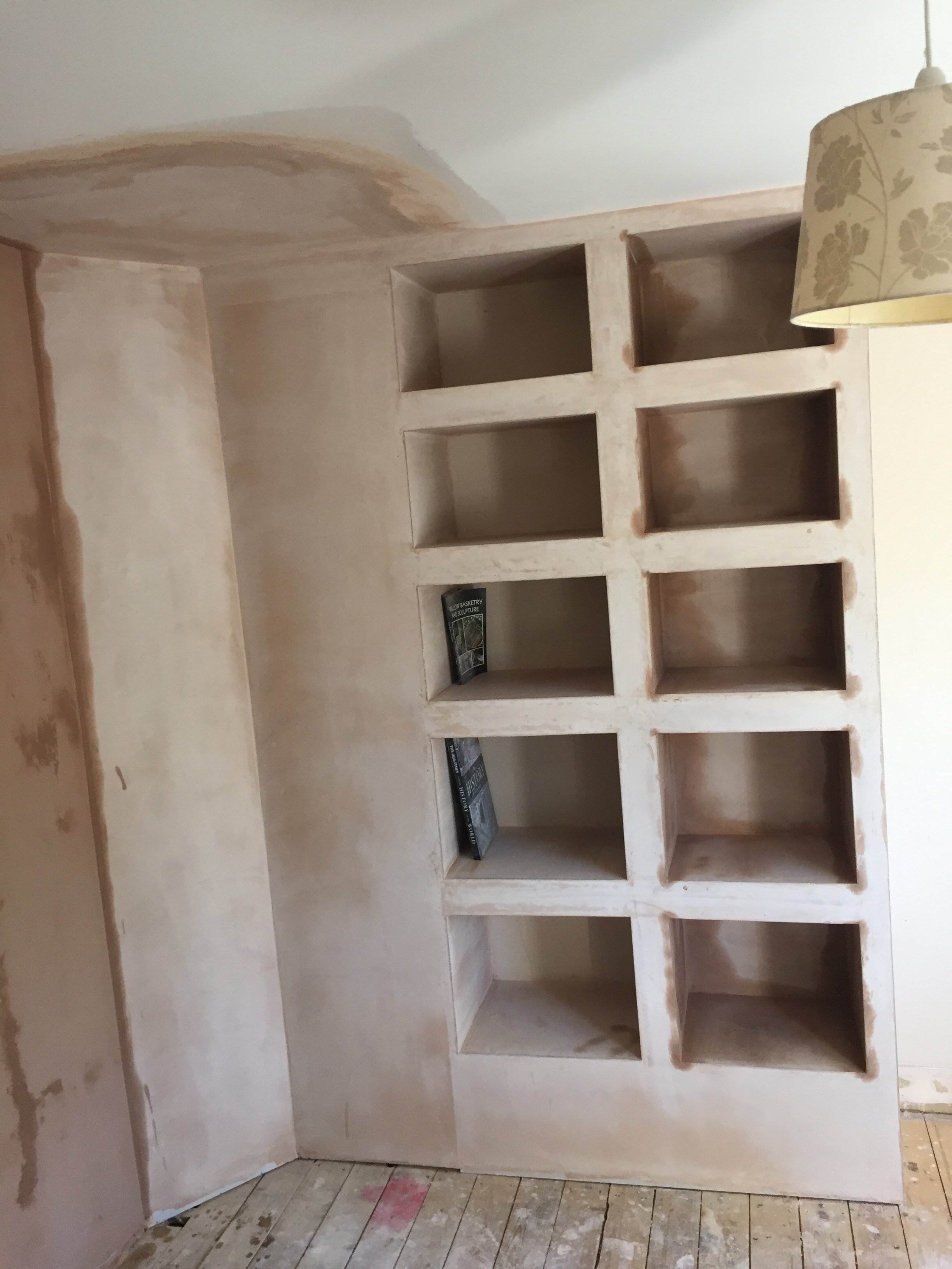 Unfinished built-in wooden bookshelf with nine empty compartments, installed against a partially painted wall.