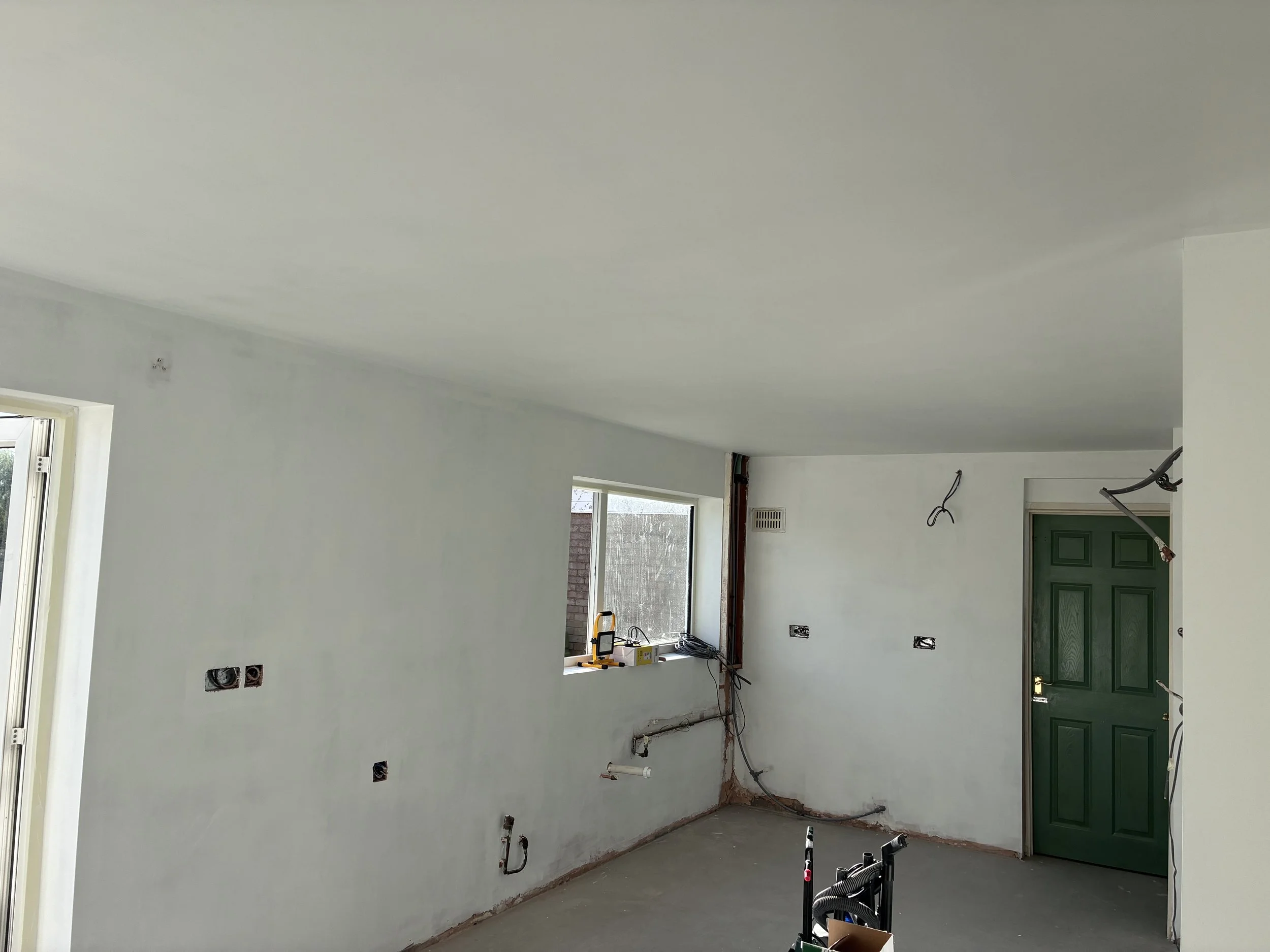 Interior of a room under renovation with exposed wiring, pipes, and outlets, a green door, and windows providing natural light.