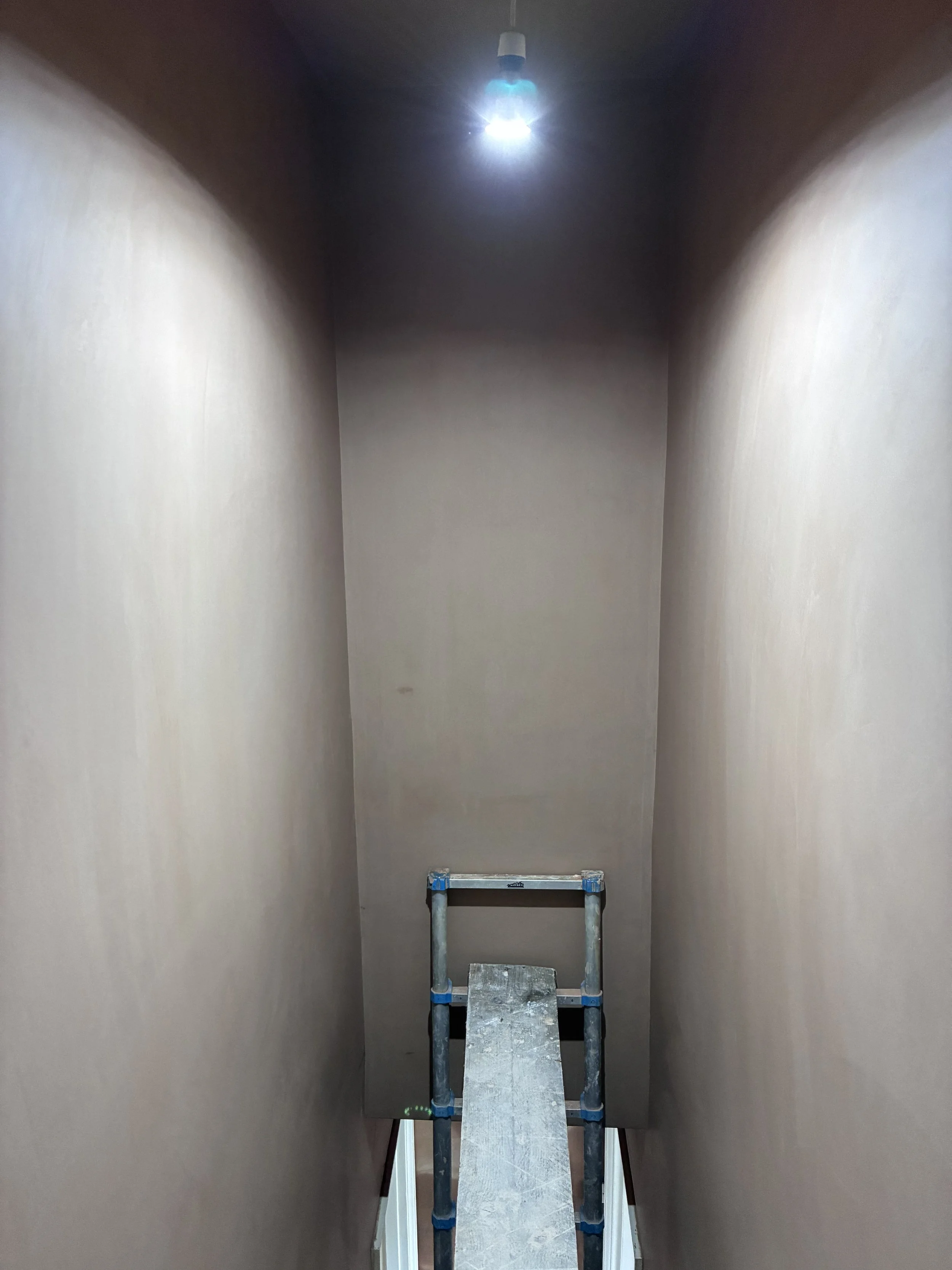 An indoor staircase with a metal scaffold placed on it, leading up to a ceiling with an exposed light fixture.