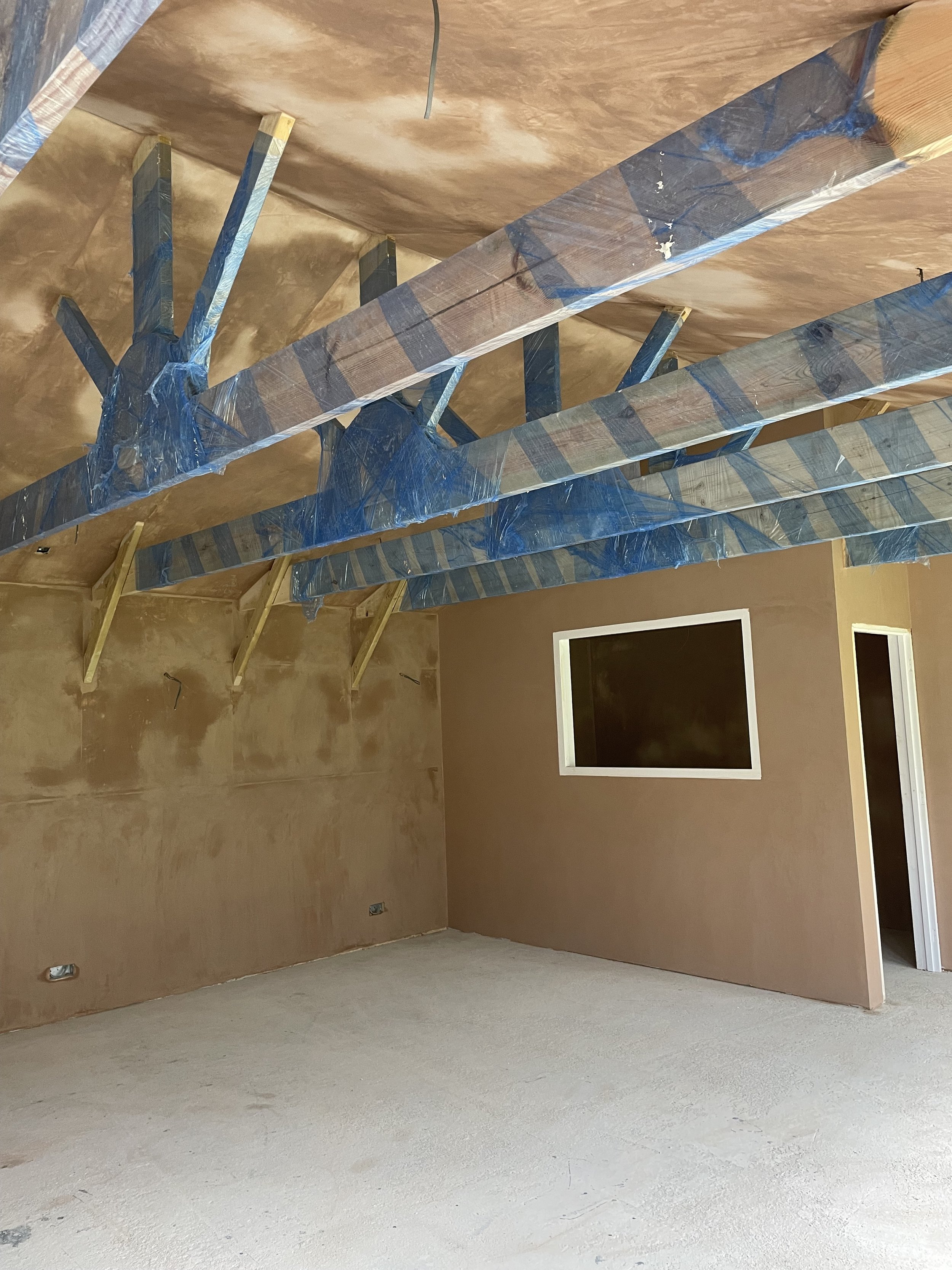 Room under construction with exposed beams and insulation wrapped in blue plastic, drywall partially installed, and unfinished walls and ceiling.