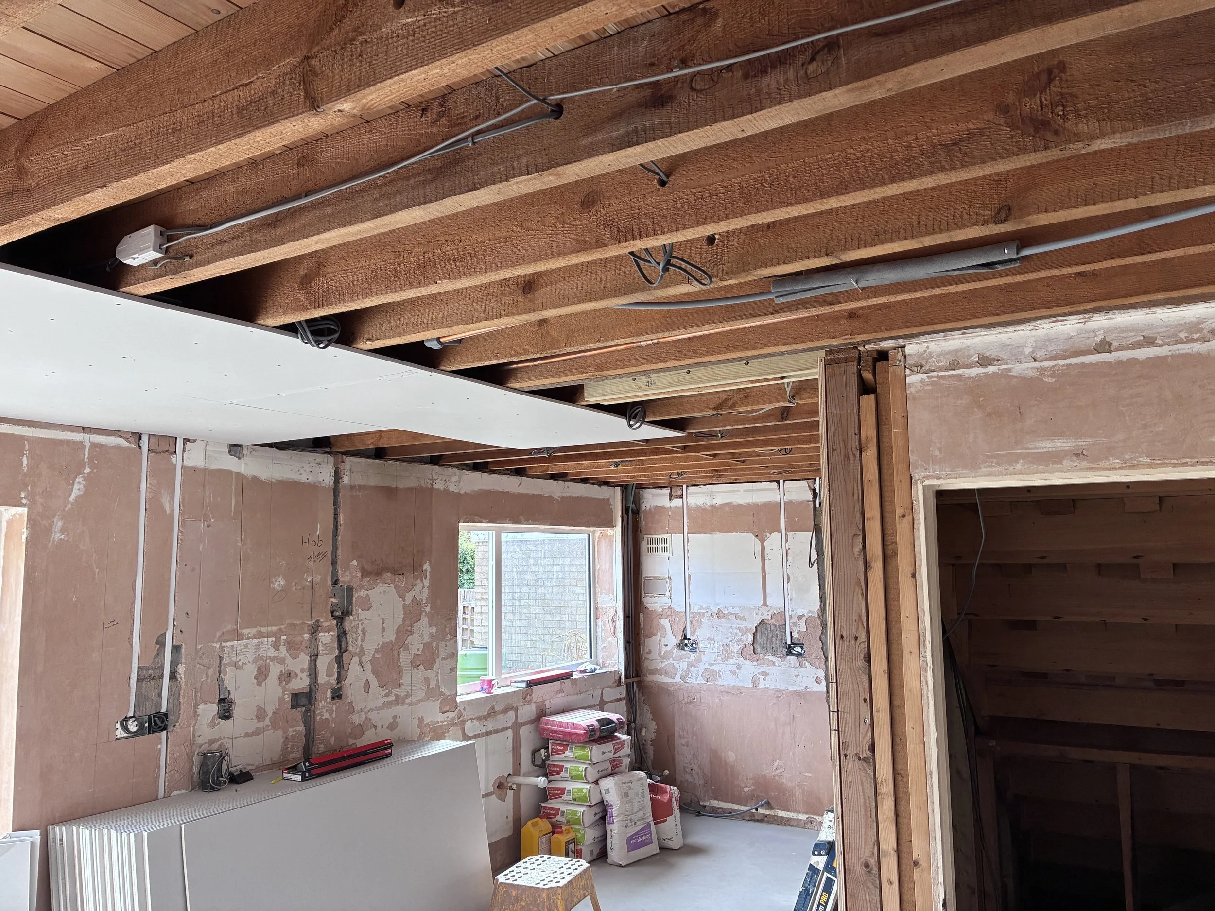 Room under construction with exposed wooden beams, electrical wiring, and construction supplies such as bags of drywall mud, held-up drywall panels, and tools.
