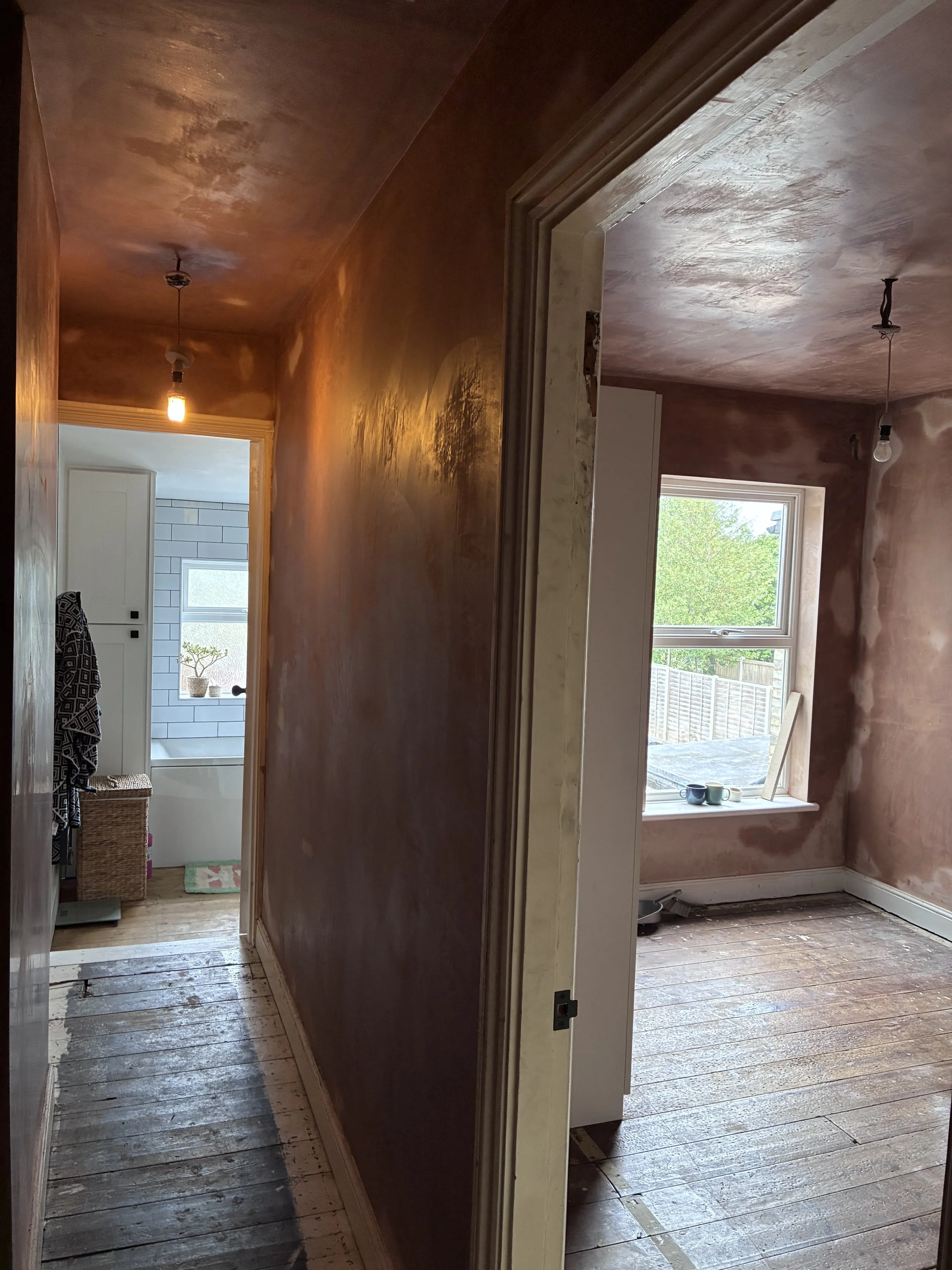 Interior of a house under renovation with hardwood floors, partially painted walls, and an open doorway leading to a room with a window and a view of green trees outside.