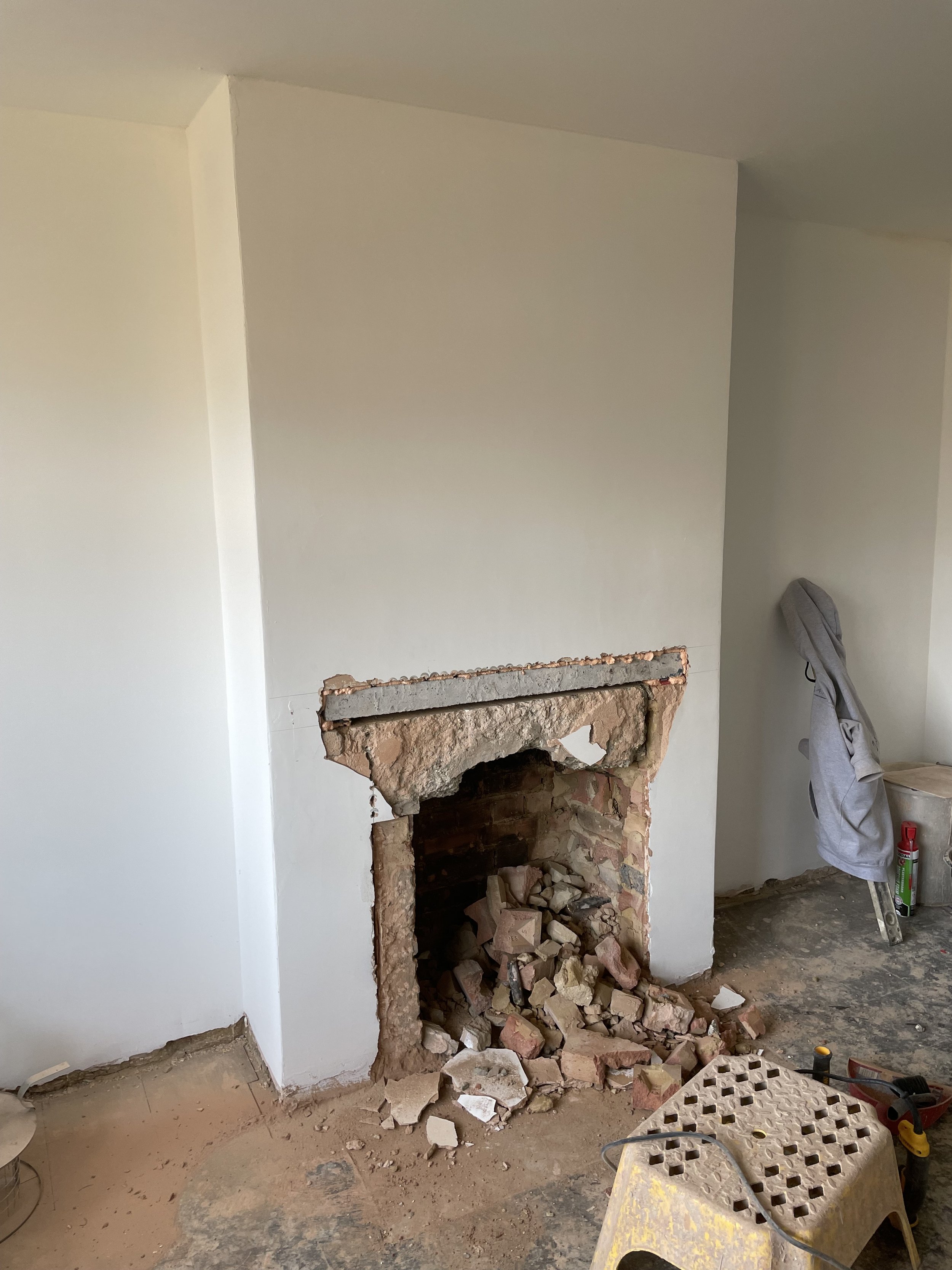 Demolition of a fireplace showing exposed brick and debris inside the hearth, with renovation tools and a chair nearby.