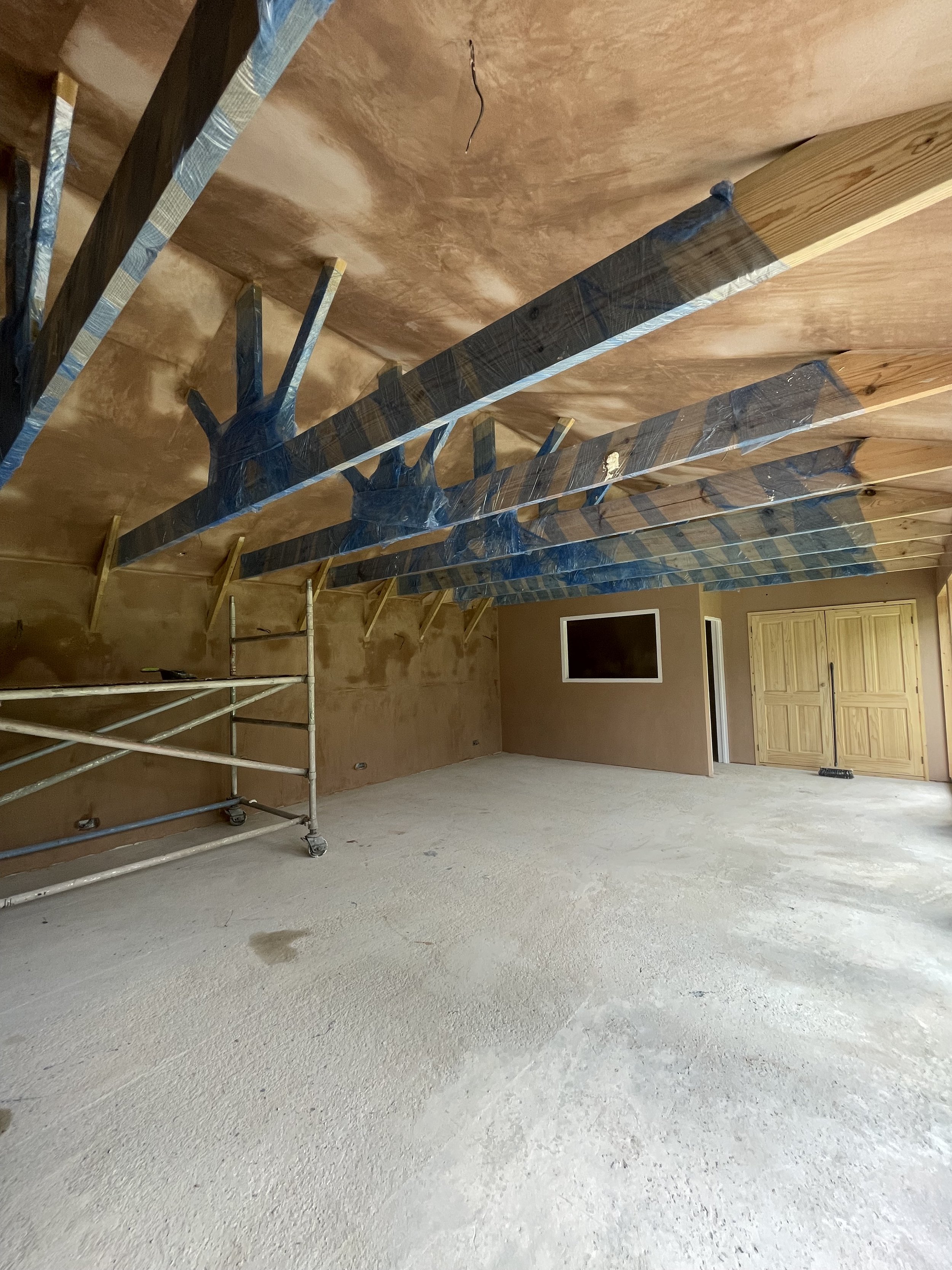 Interior of a room under construction, with exposed wooden beams on the ceiling wrapped in plastic, a scaffolding on the left, and unfinished walls and flooring.