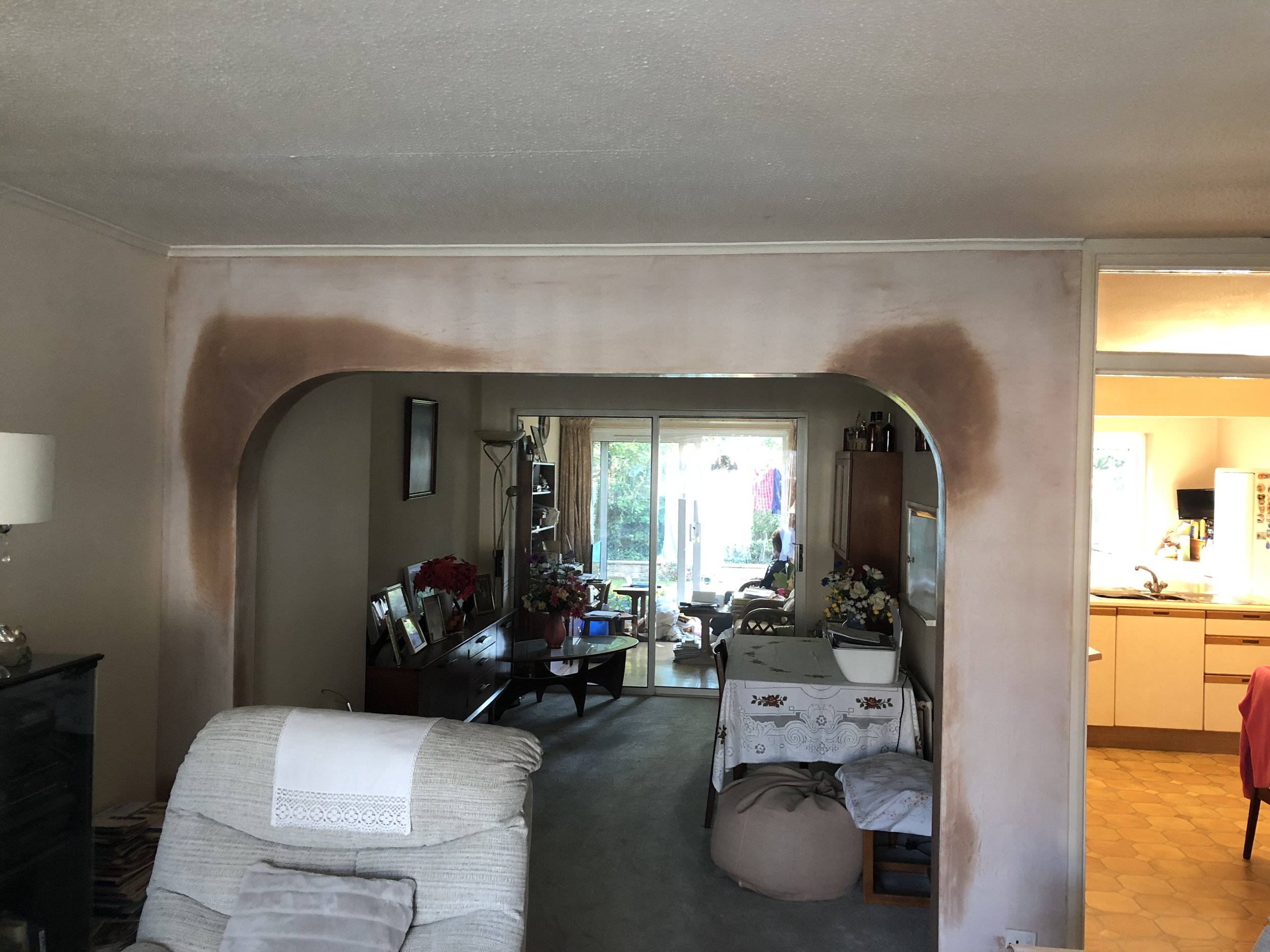 Living room with an arched opening, a white armchair with a towel on it, a side table, a dining table with a tablecloth, and a view into a kitchen with yellow cabinets and a person sitting in the background.