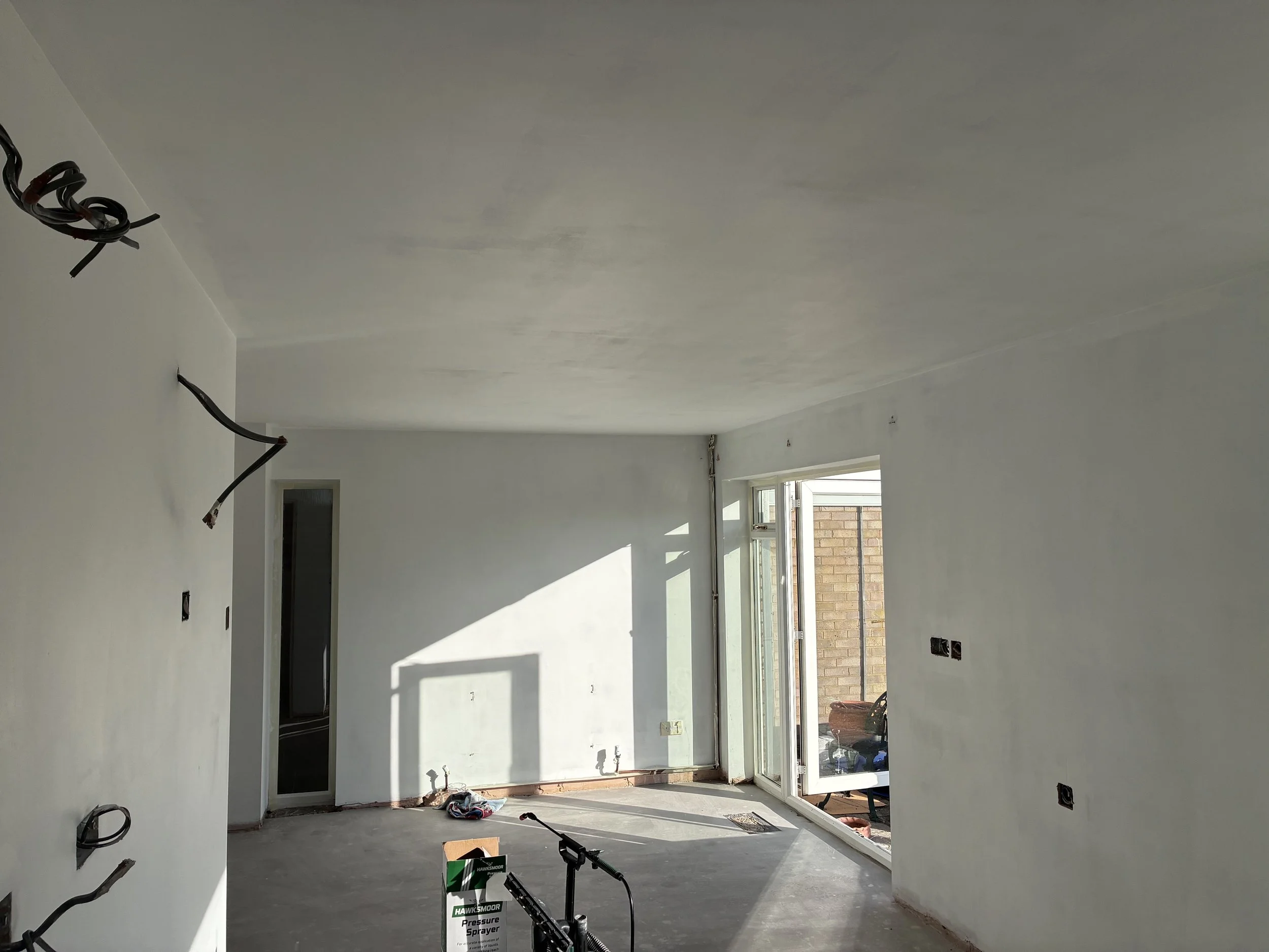 A room under construction with unfinished walls and ceiling. Exposed electrical wiring and outlets, large window, and construction tools on the floor.