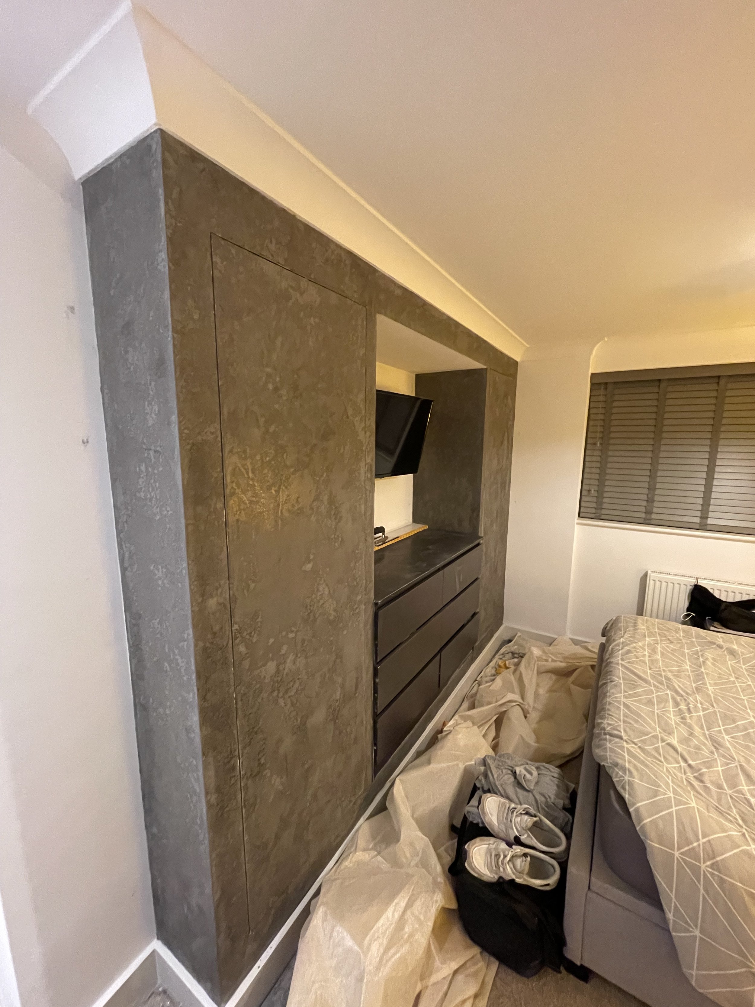 Unfinished bedroom with a custom wall feature, a bed with a patterned duvet, suitcase with shoes, and a window with closed blinds.