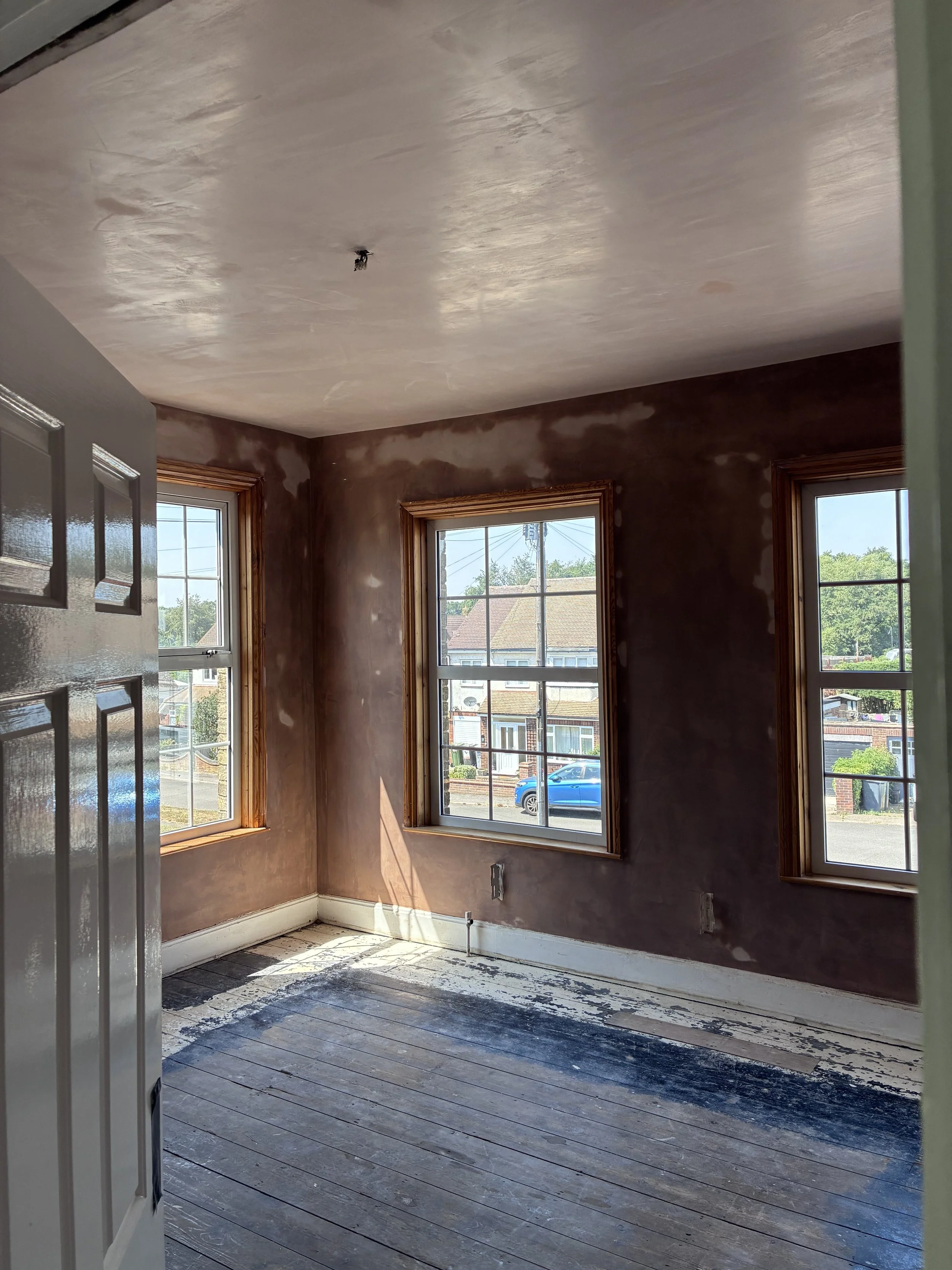 An empty room under renovation with three windows showing a view of houses and trees outside. The walls are partially finished with patches of drywall, and the floor is stripped, revealing worn wood planks with paint residue. There is a ceiling light fixture hole but no light installed yet.