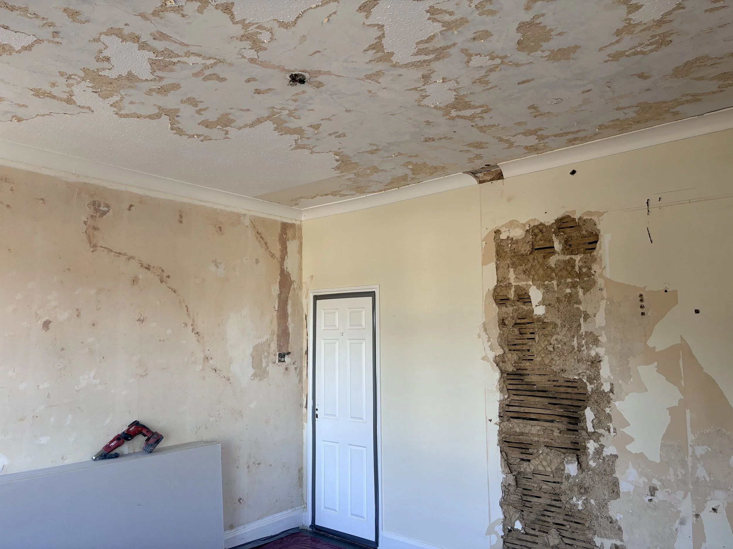 A room undergoing renovation with peeled wallpaper and exposed drywall, a white door, and construction tools on the floor.