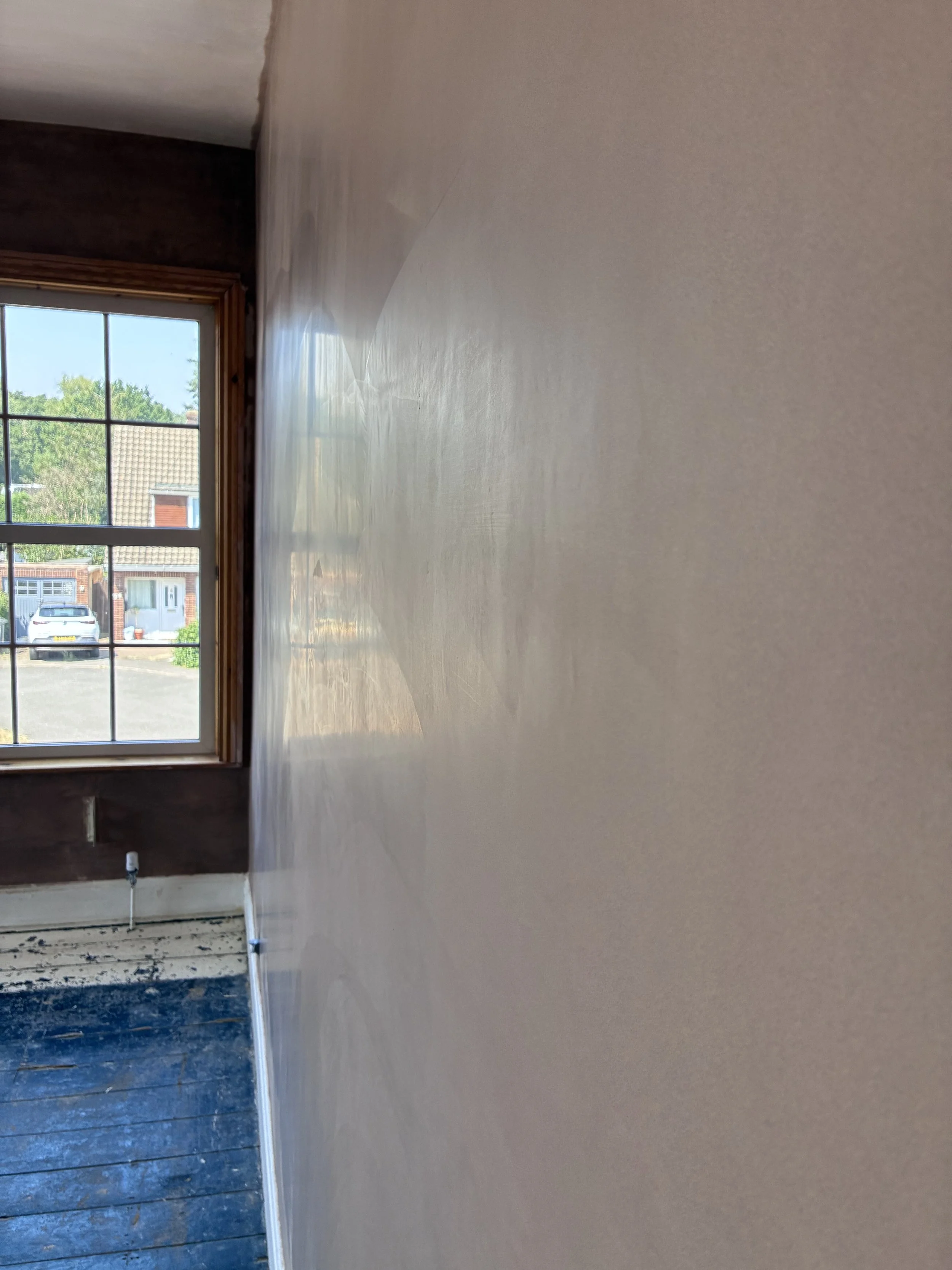 Interior view of a room with a window showing trees and houses outside, walls partially painted with a light beige/peach color, and part of the floor and baseboard visible, with some construction or renovation work in progress.