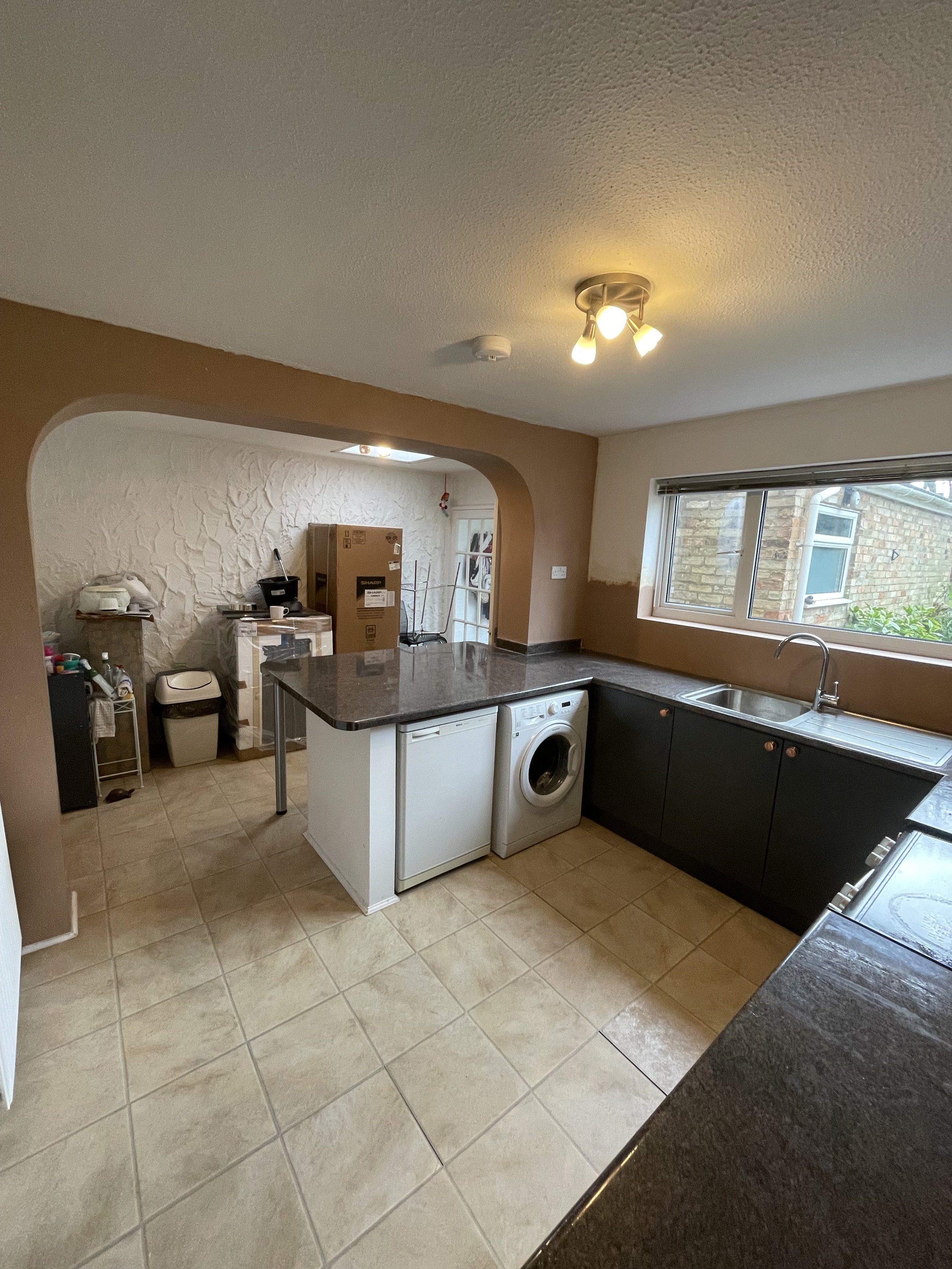 Kitchen with black cabinets, a stainless steel sink by a large window, a washing machine, and cardboard boxes in the background separating the kitchen from another room.