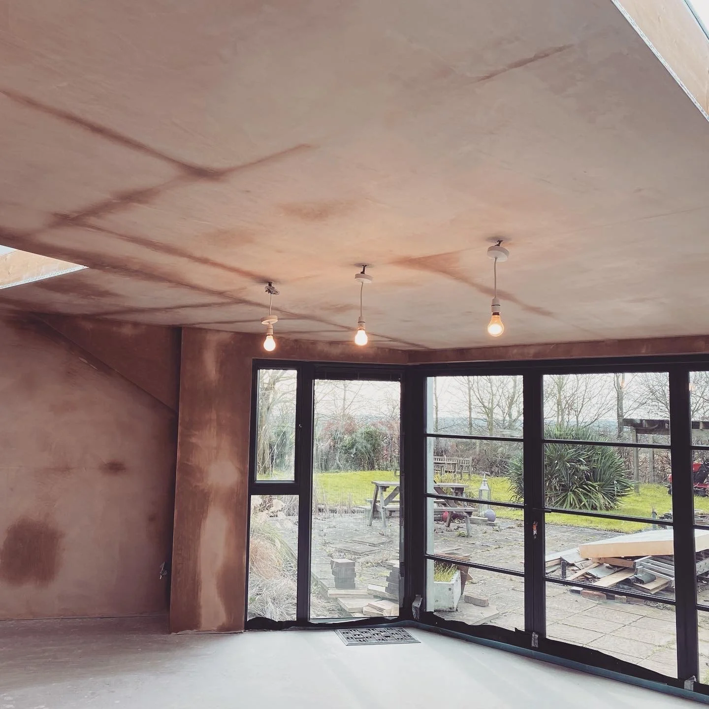 Interior of a room under construction with unfinished wooden walls and ceiling, three hanging light bulbs, large sliding glass doors, and an outdoor garden area with trees, plants, and outdoor furniture.