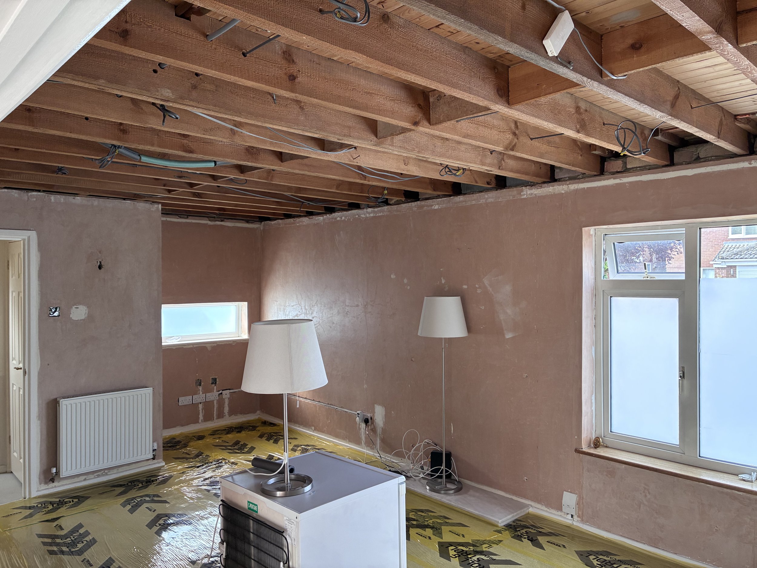 Living room under renovation with exposed wooden ceiling beams, unfinished walls, a heater, a standing lamp, and a window showing a brick building outside.