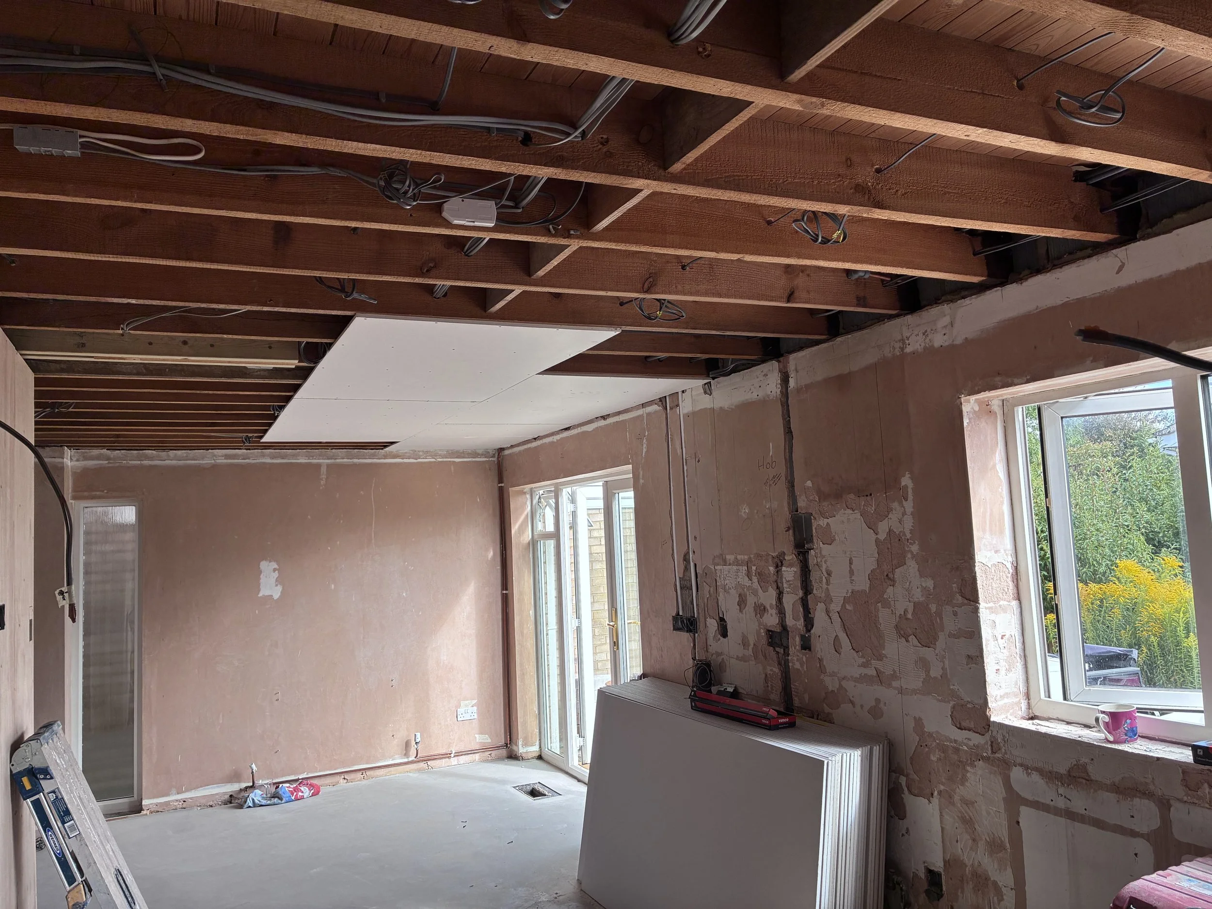 Refurbishment in progress: construction room with exposed wooden ceiling framework, electrical wiring, unfinished walls, and a window revealing an outdoor view with greenery.
