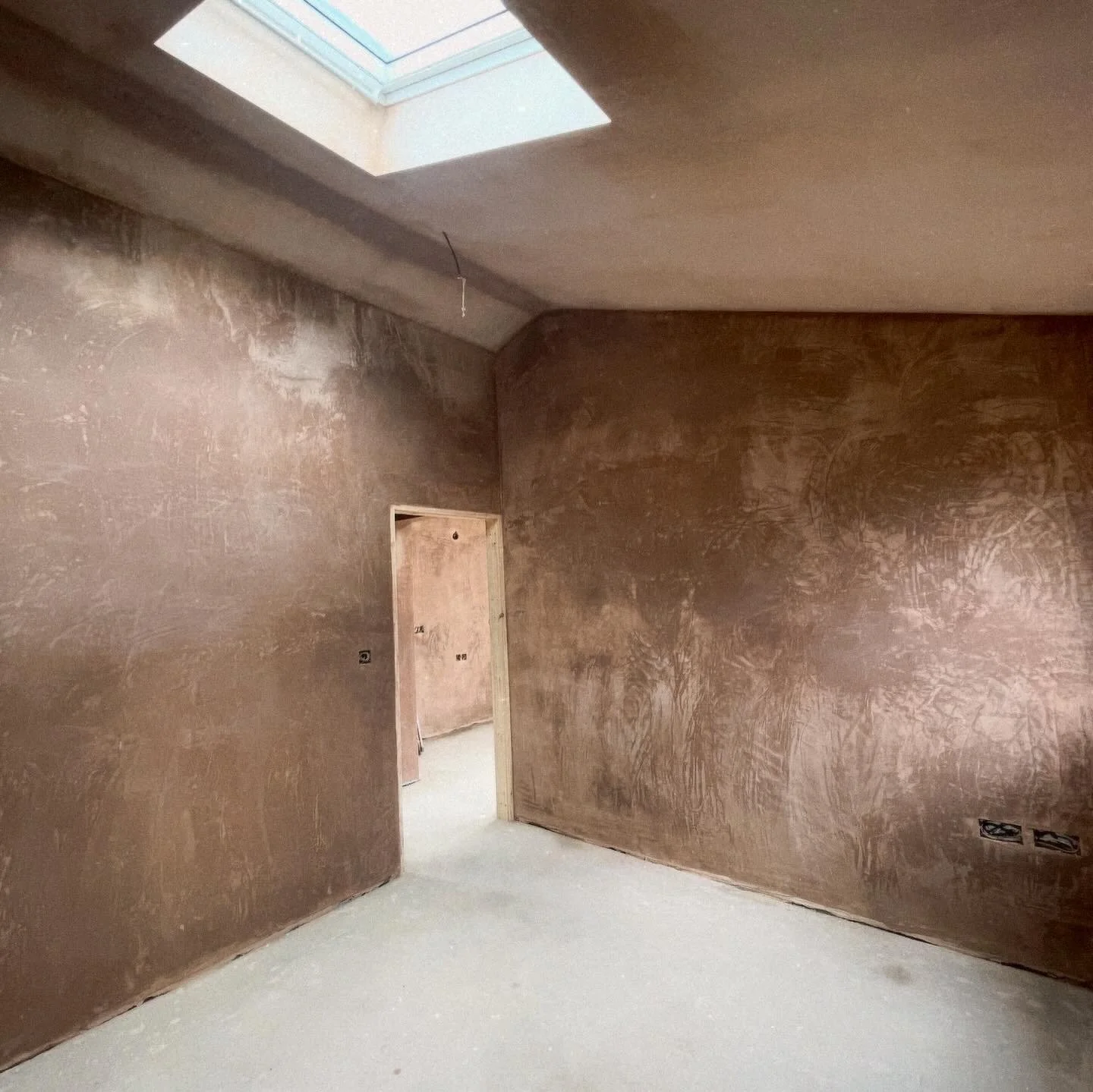 An unfinished room with brown plastered walls, a window in the ceiling, and an open doorway leading to another room. The floor appears uncarpeted or unfinished.