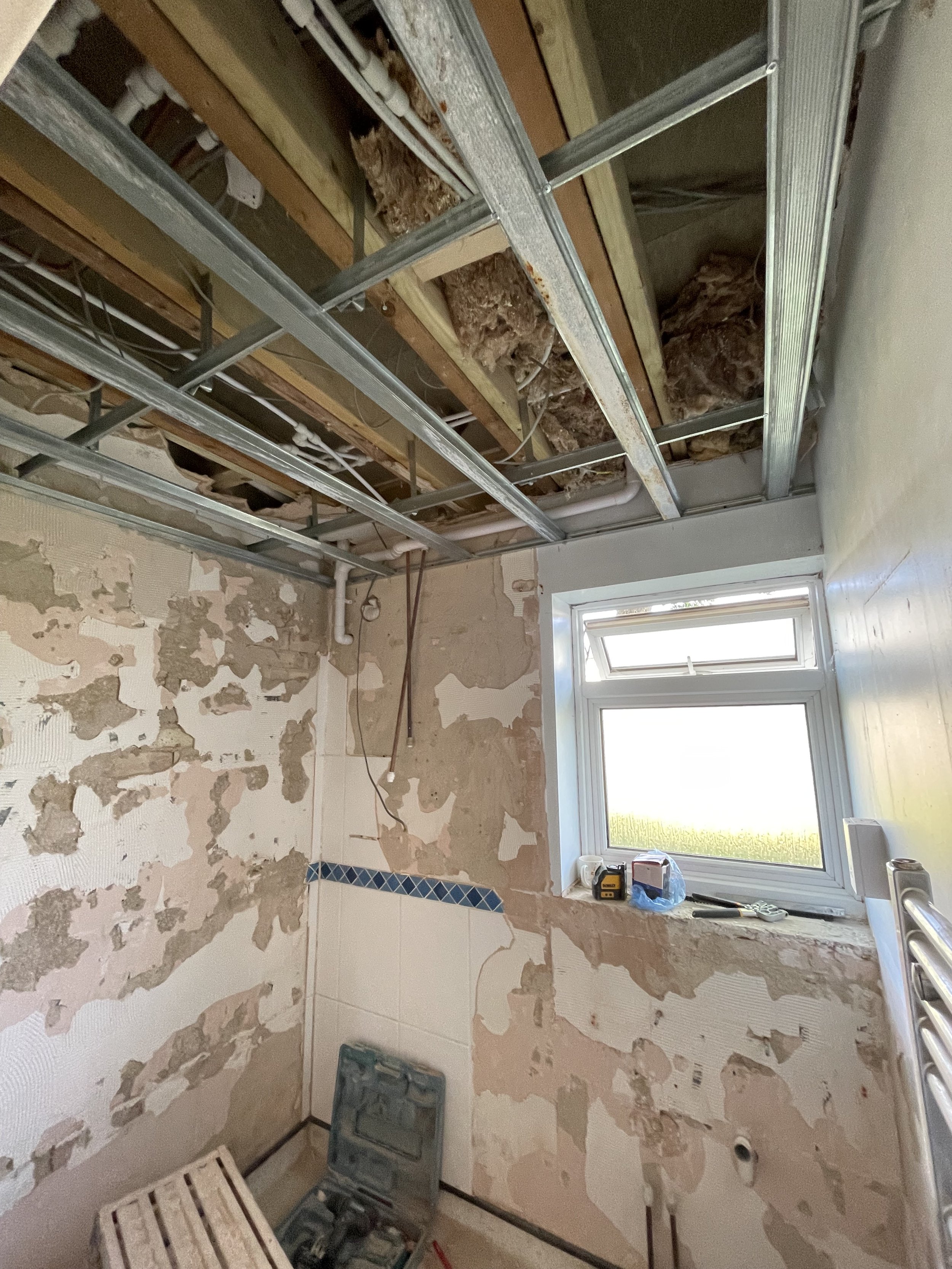 A room under renovation with exposed wall studs and insulation in the ceiling. The wall has partially removed wallpaper revealing drywall and tile. A window with items on its sill, including a toolbox, tape measure, and a roll of paper. Construction 