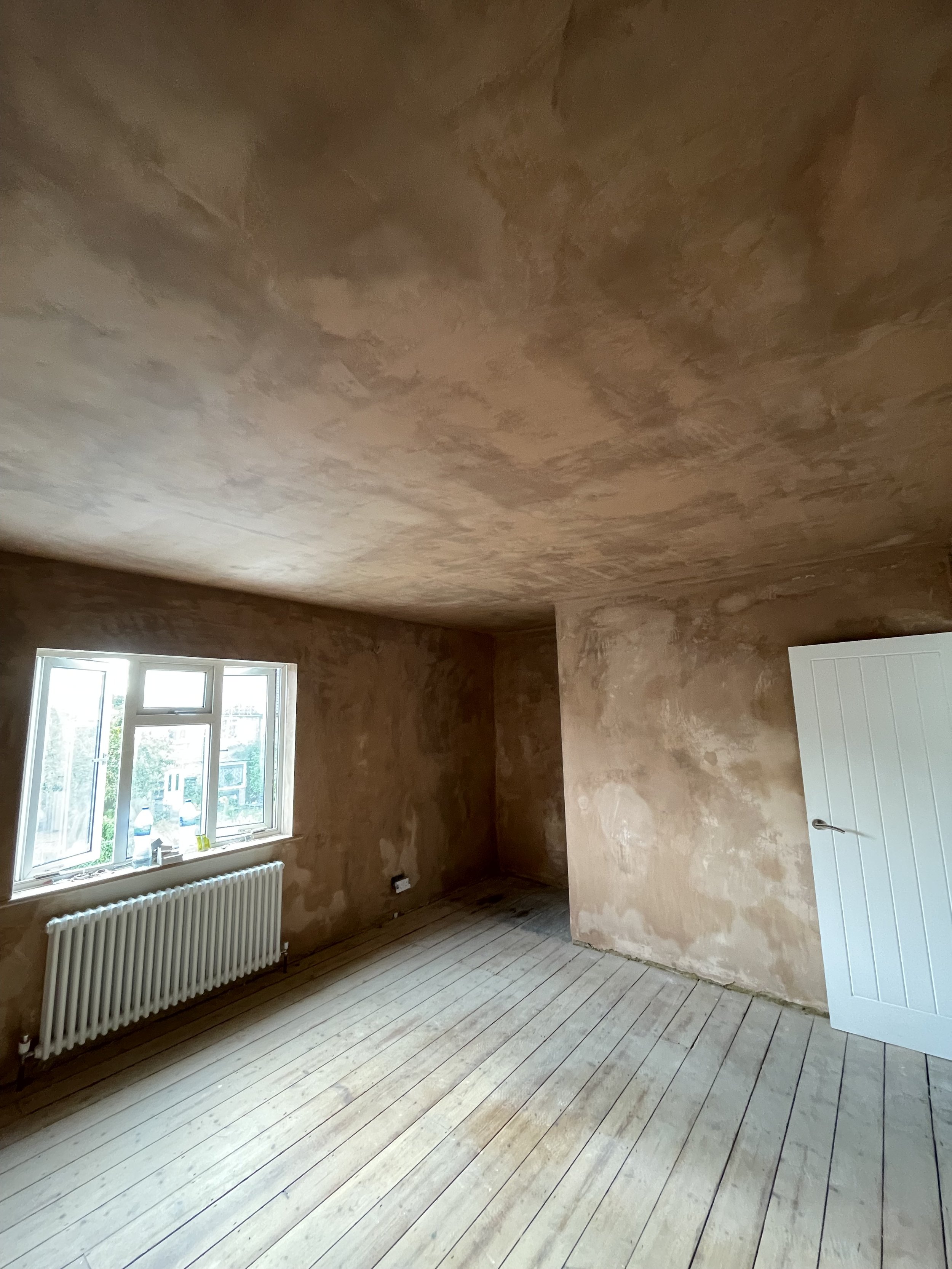 An empty room with unfinished beige walls and ceiling, a window with a white radiator below it, and a white door on the right side.