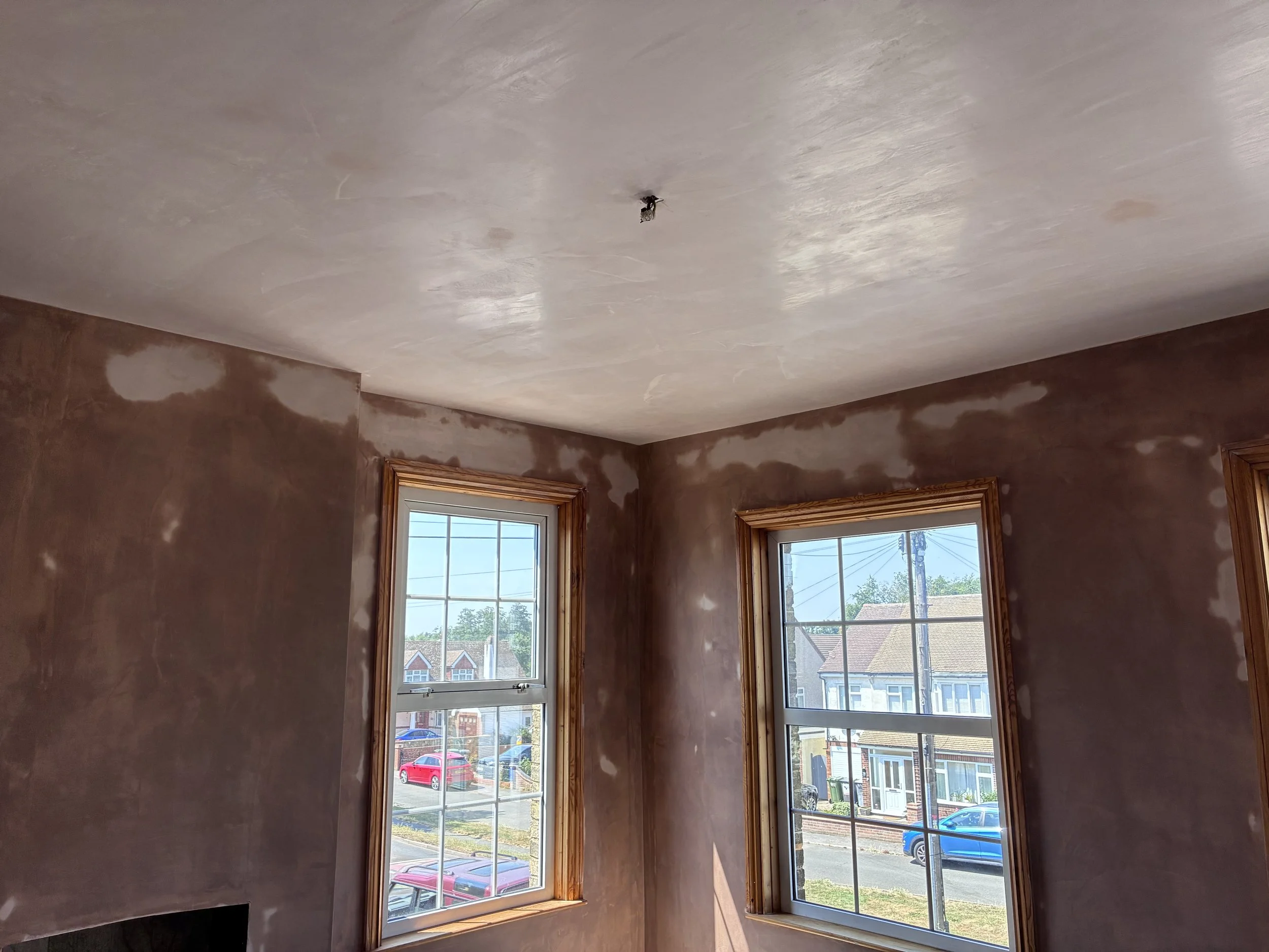 A room with two windows showing a neighborhood outside, and a ceiling with a small electrical fixture from which wires hang.