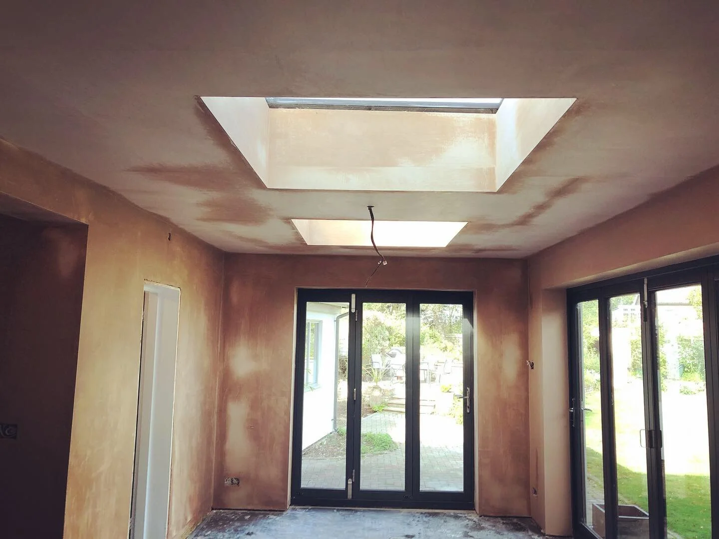 Room with large sliding glass doors leading to outdoor patio, unfinished walls and ceiling with two skylights, construction wires hanging from ceiling.