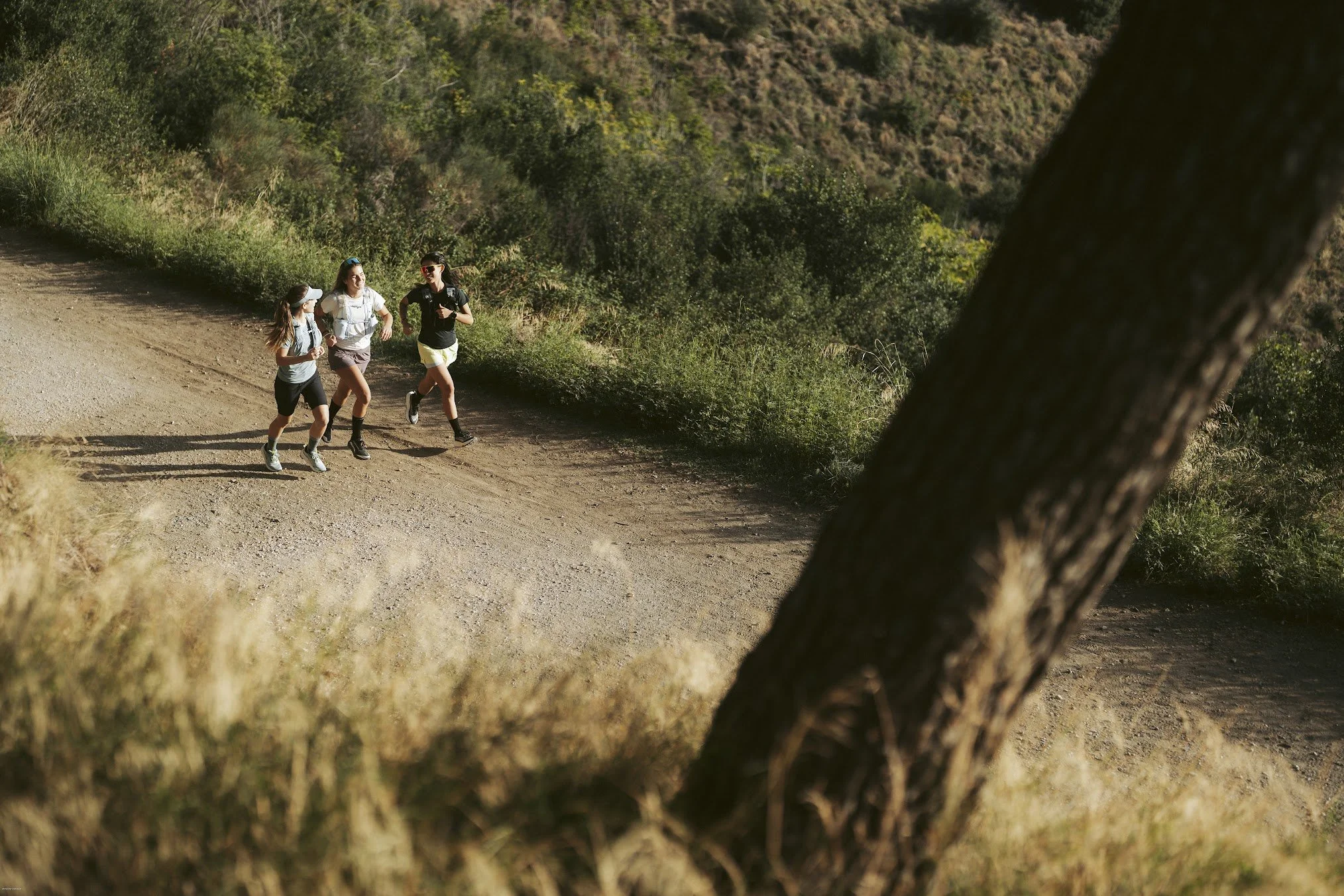 Three women are running on a dirt trail in a hilly, wooded area during daytime. They are dressed in athletic clothing and are wearing sunglasses.