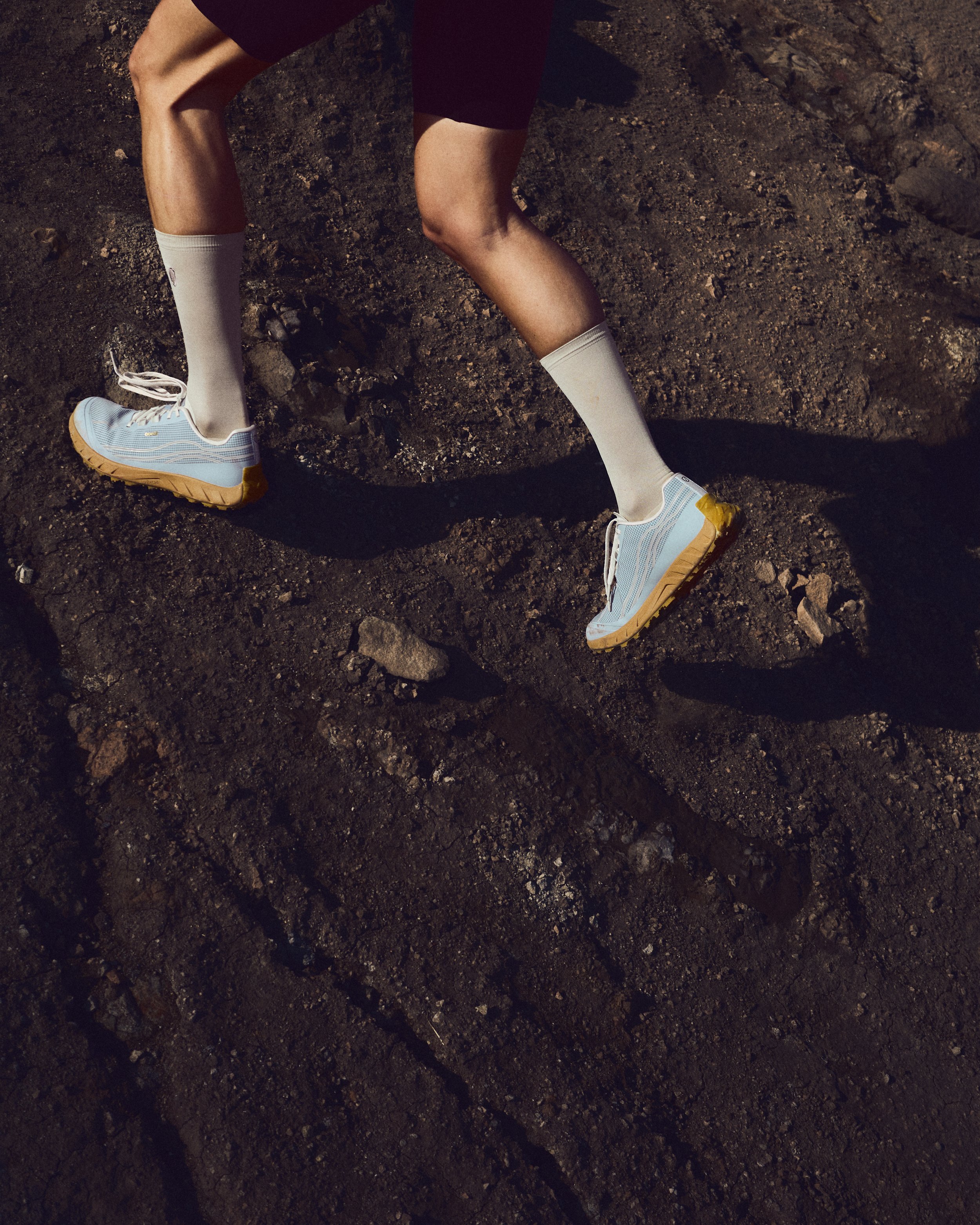 Close-up of a person's legs wearing blue and yellow athletic shoes, white knee-high socks, and black shorts. The person appears to be standing or walking on dark, rocky dirt ground. Norda.