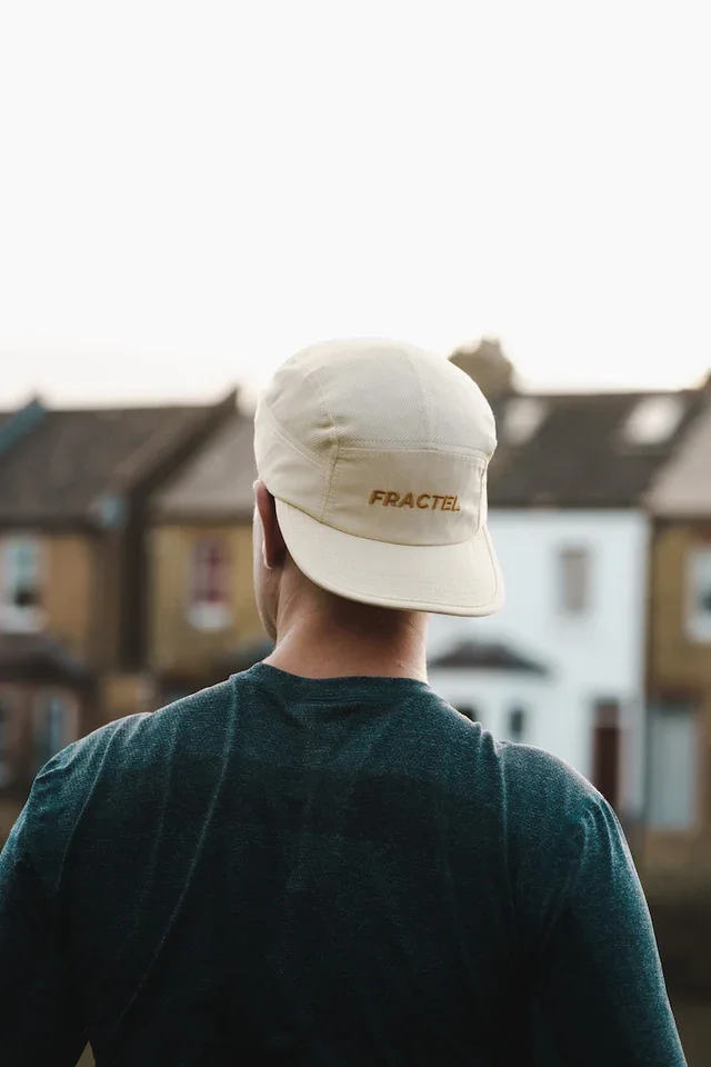 Back view of a person wearing a beige cap with the word 'FRACTEL' embroidered on it, standing outdoors with houses in the background.