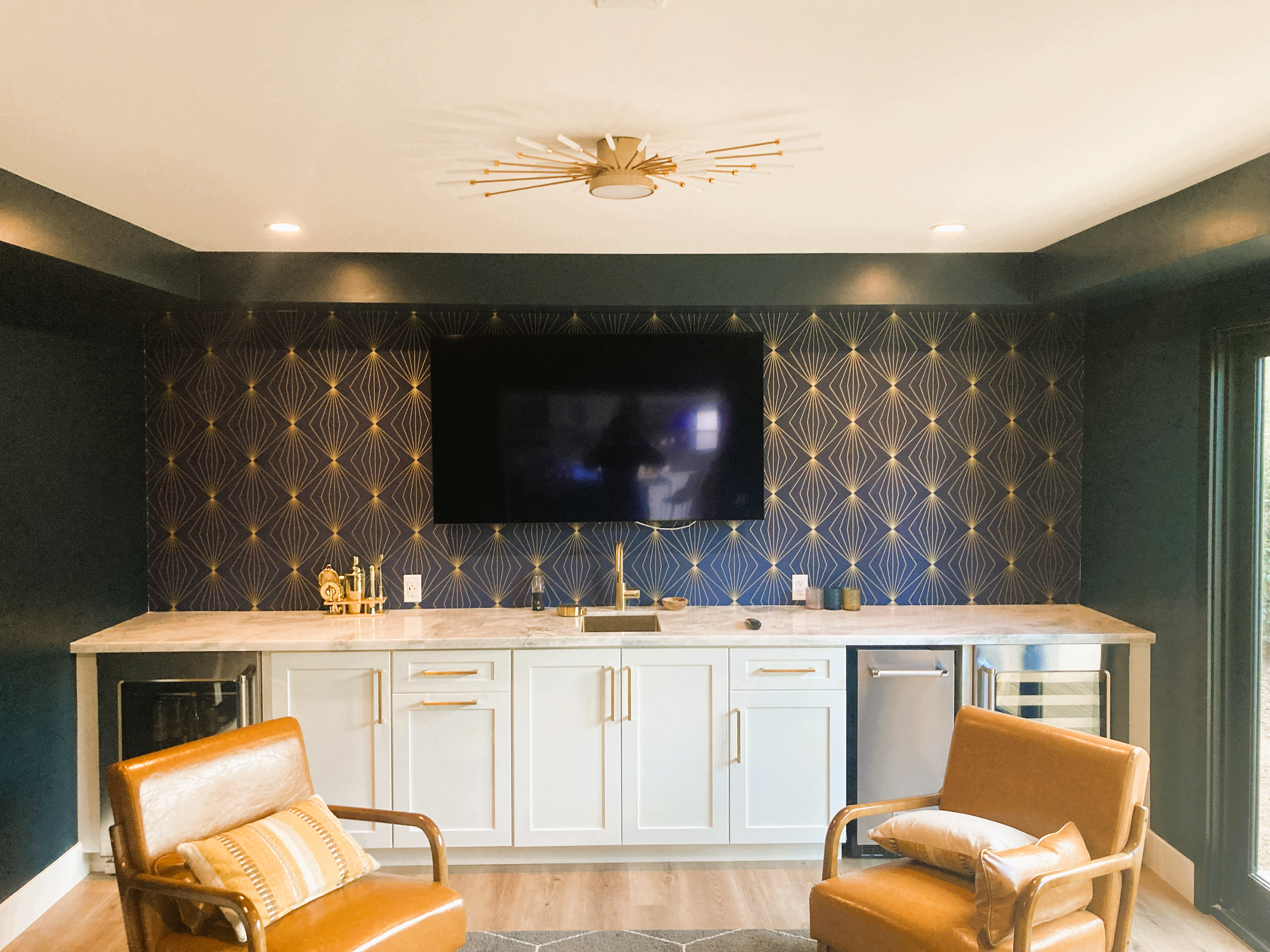 Modern kitchen with a marble countertop, gold accents, a large flat-screen TV wall, and two leather chairs with pillows.