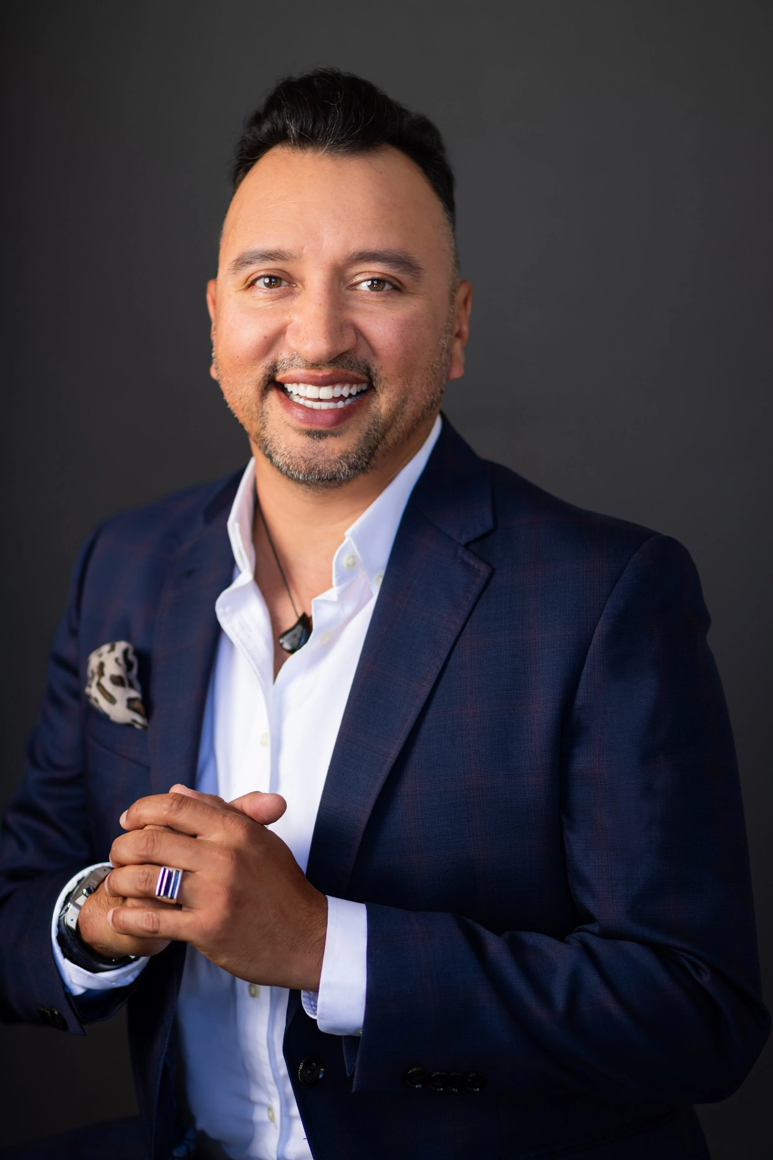 A smiling man with short dark hair, wearing a navy blue suit, white shirt, and a patterned pocket square, standing with hands clasped against a dark background.