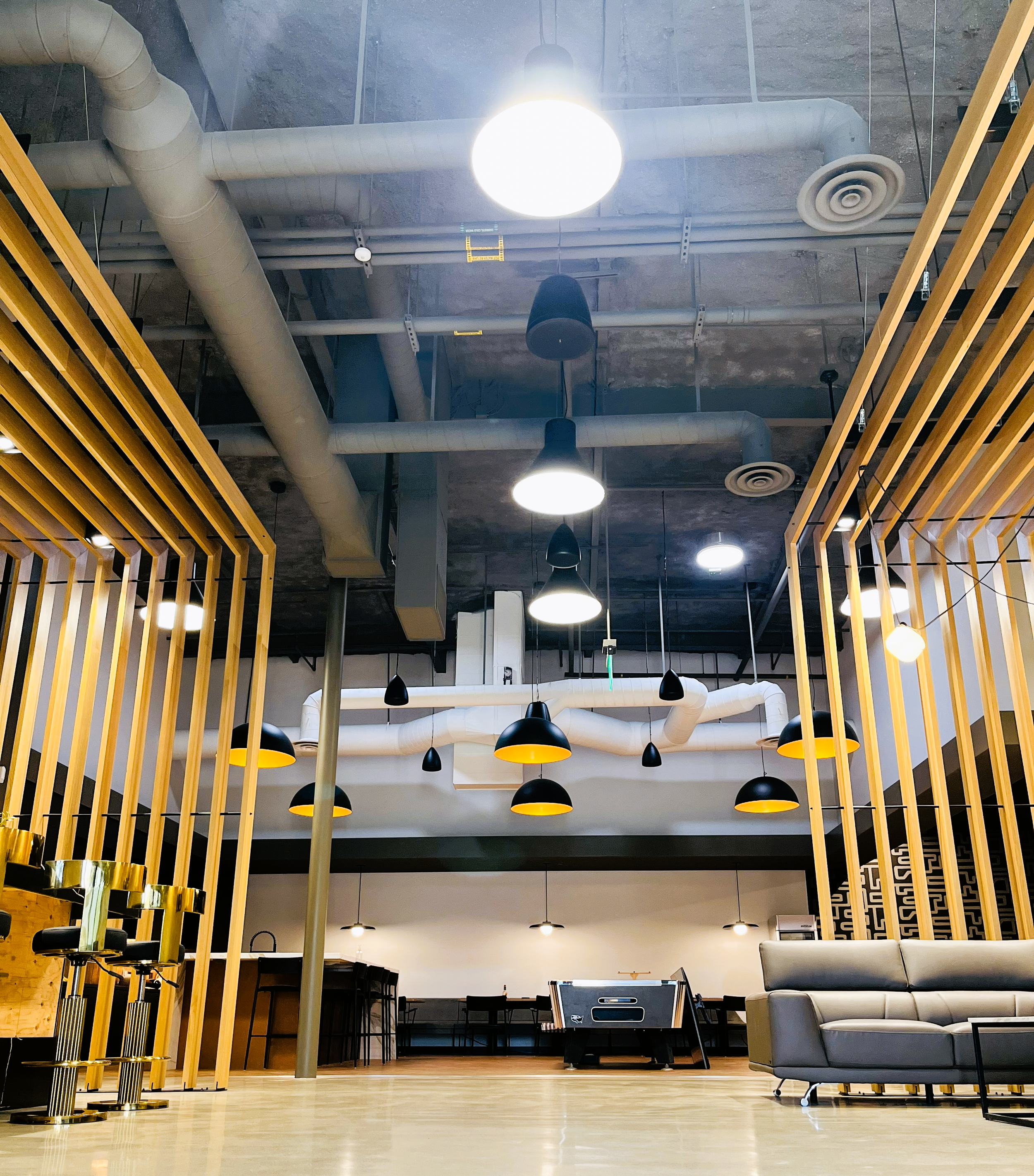 Interior of a modern office or lounge area with exposed ceiling pipes, multiple hanging black and white ceiling lights, wooden slat partitions, a gray sofa, and a foosball table.