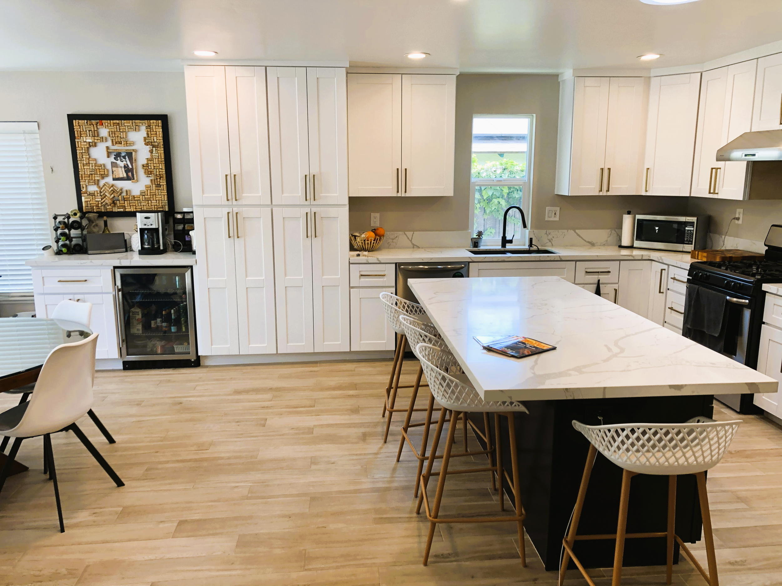 Modern kitchen with white cabinets, a central island with a white marble countertop, wooden bar stools, a black stove, microwave, and a farmhouse sink, with hardwood flooring and natural light from a window.