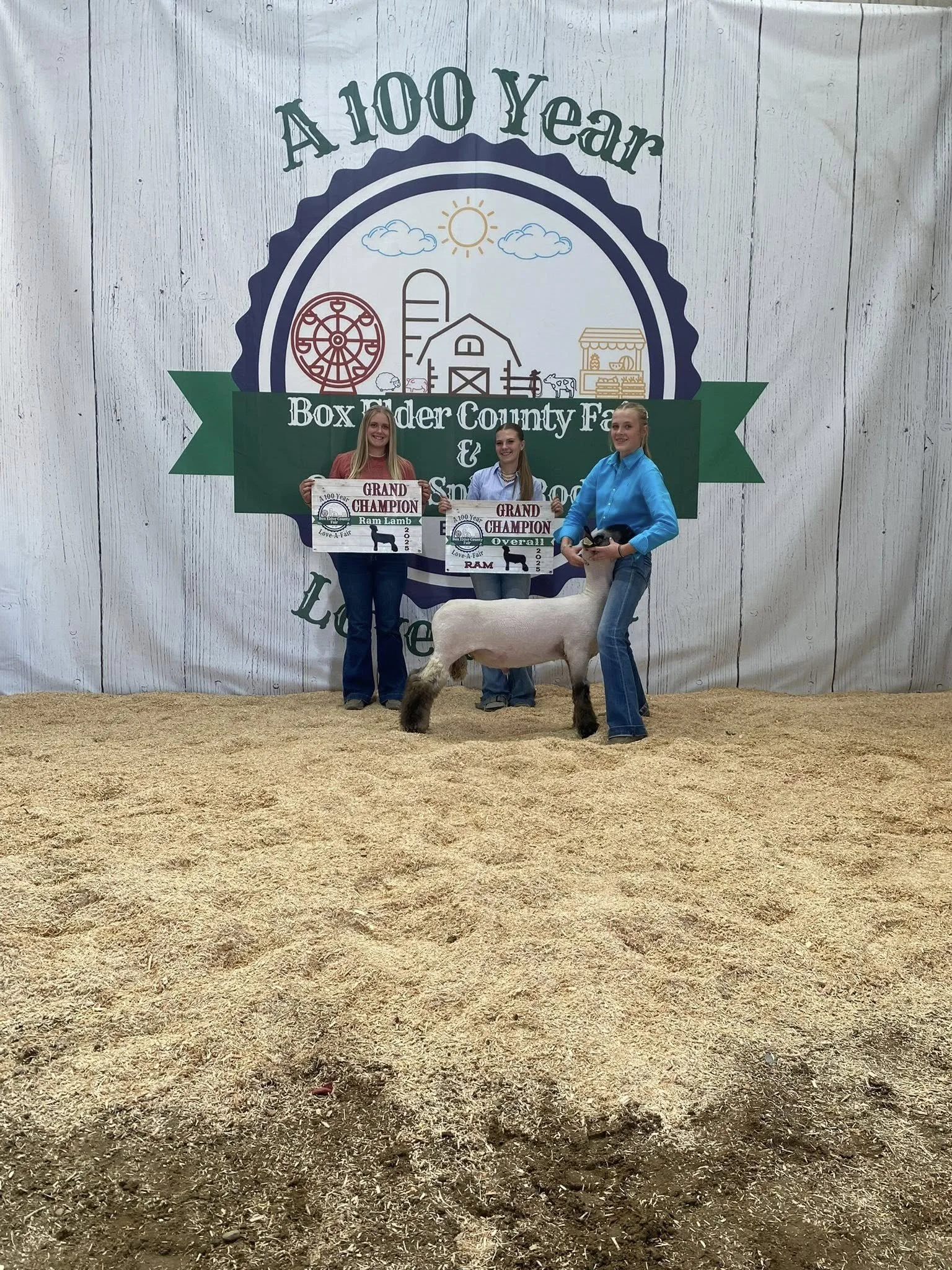 Three young women standing on a dirt floor in front of a large banner celebrating a 100-year event, holding award signs and posing with a sheep.