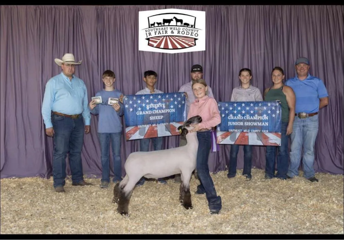 A group of eight people on stage at the Southeast Weld County Jr Fair & Rodeo, with a purple curtain backdrop. They are holding two banners that read 'Reserve Grand Champion Sheep' and 'Grand Champion Junior Showman.' A young girl in front is holding a sheep, and several people are wearing cowboy hats and casual western attire.