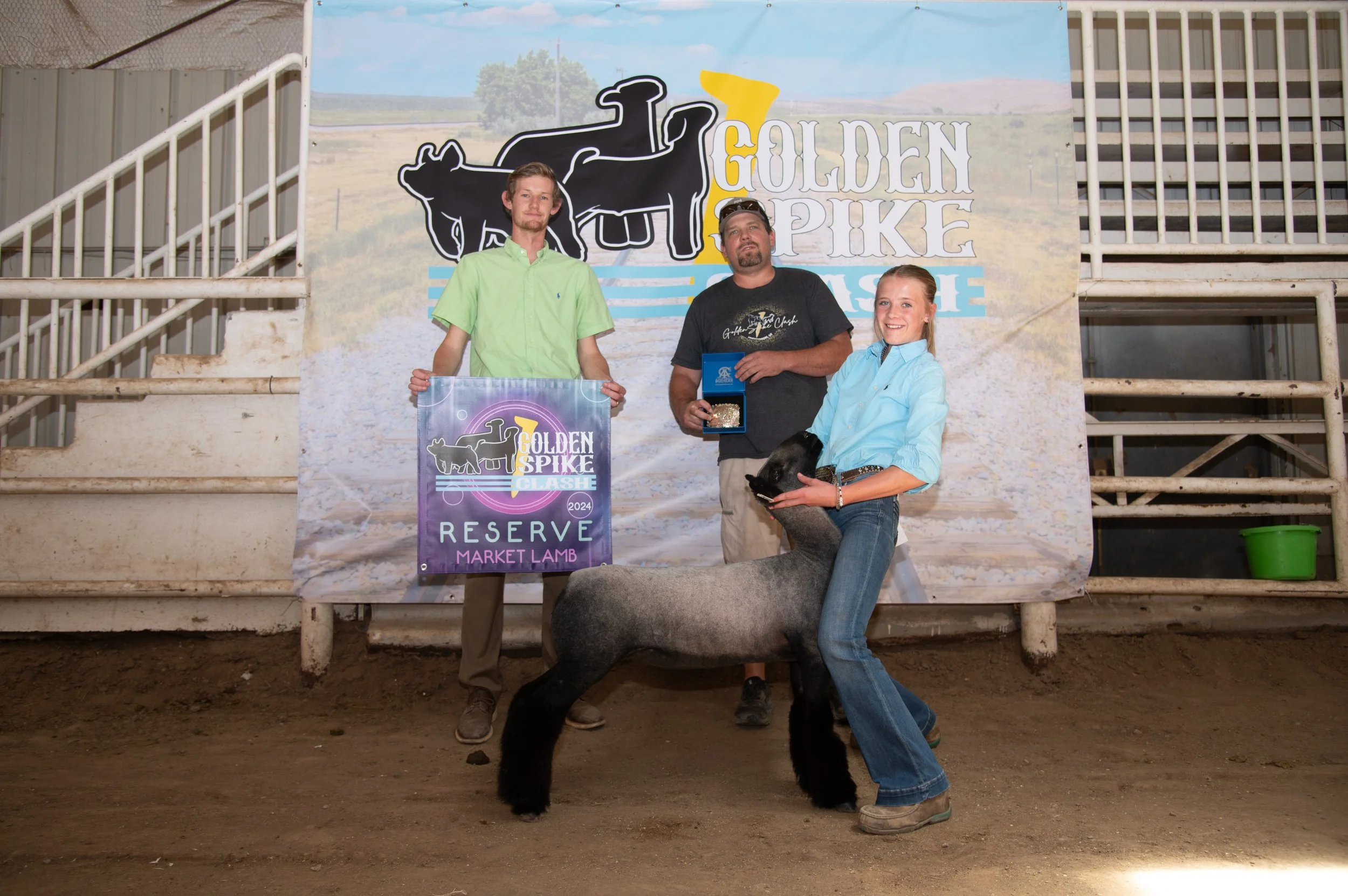 Three people standing in front of a banner at a livestock event, with a young woman holding a sheep, a man holding a medal, and a young man holding a purple prize banner that reads "Reserve Market Lamb."